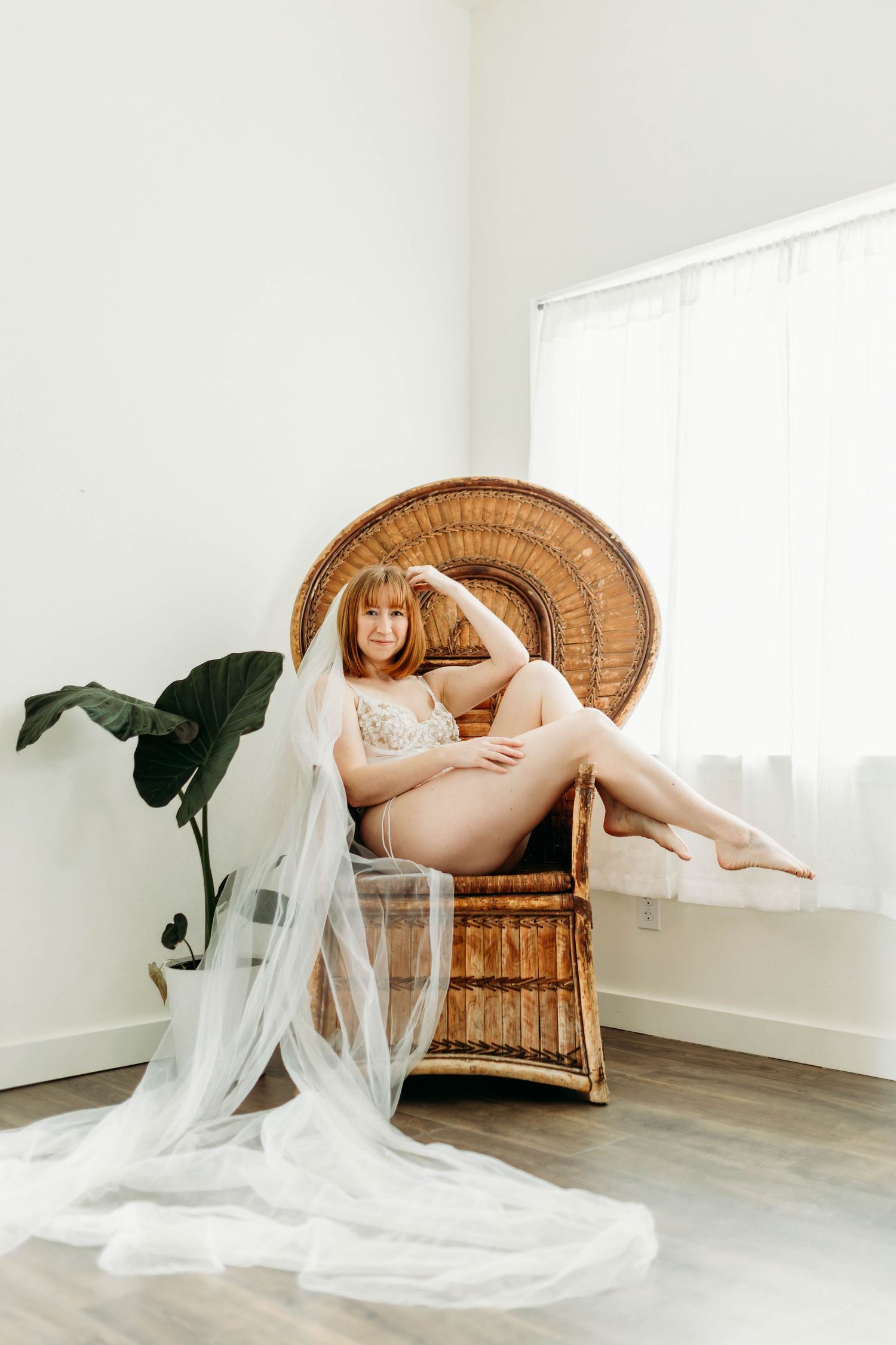 A romantic boudoir photo featuring a flowing white veil and vintage wicker peacock chair against a minimalist white wall.