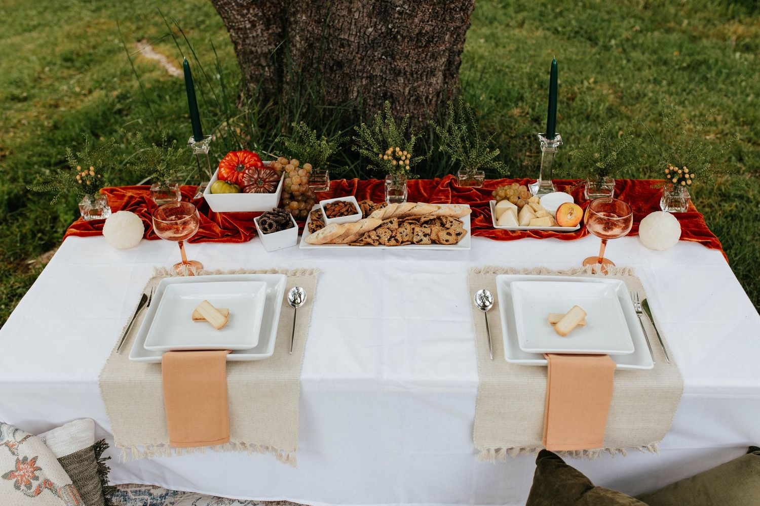 Elegant outdoor table setting with white linens, peach napkins and fall decor beneath a large tree.