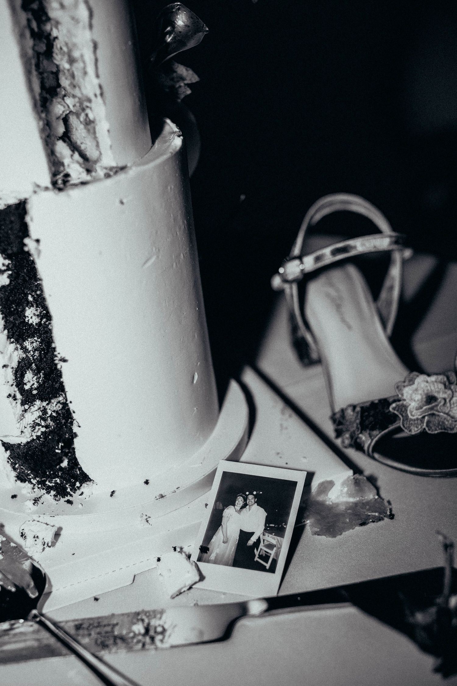 Black and white flat lay of wedding cake, high heel shoe, and vintage Polaroid photo on table.