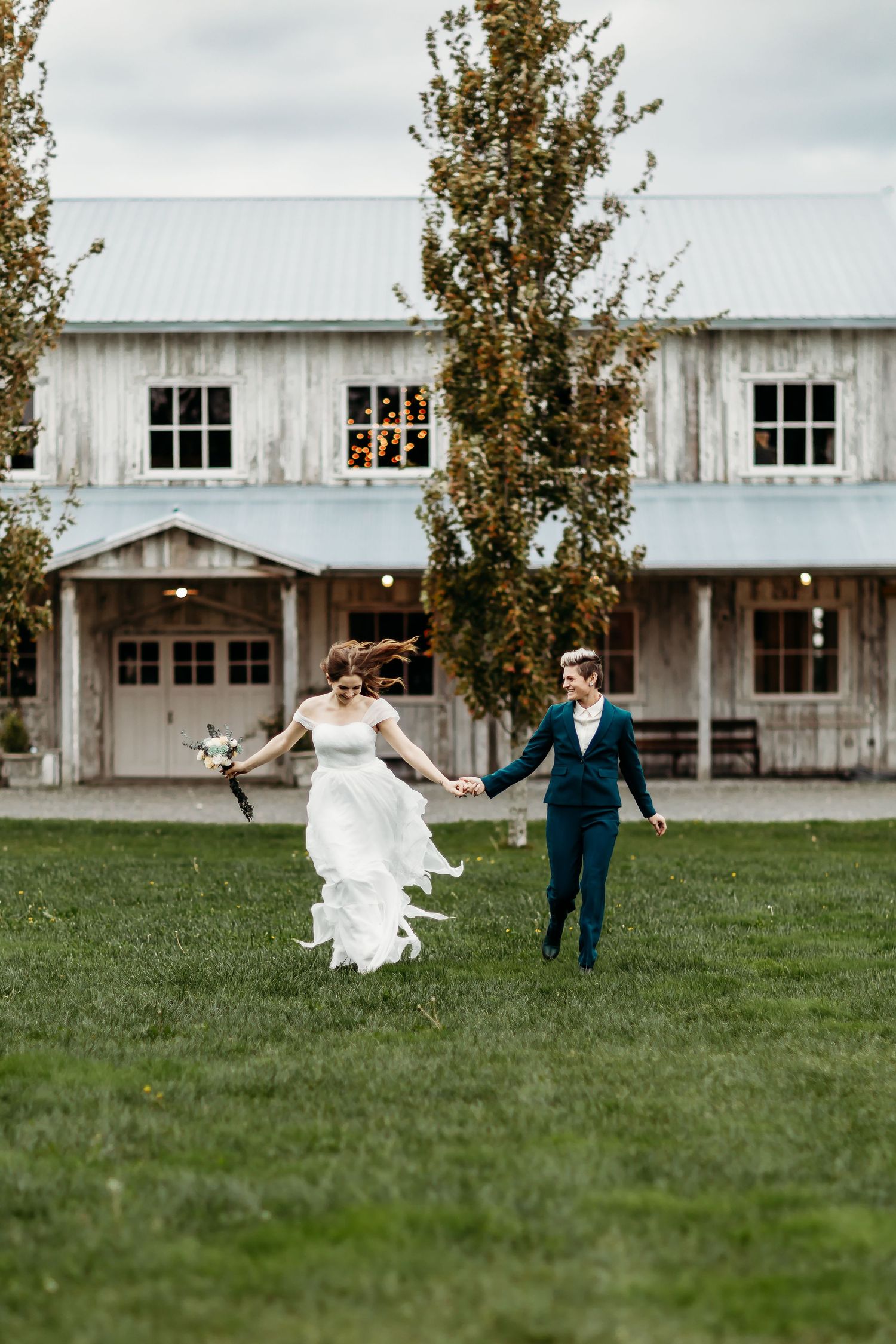 Joyful couple running on grass in front of rustic white barn building.