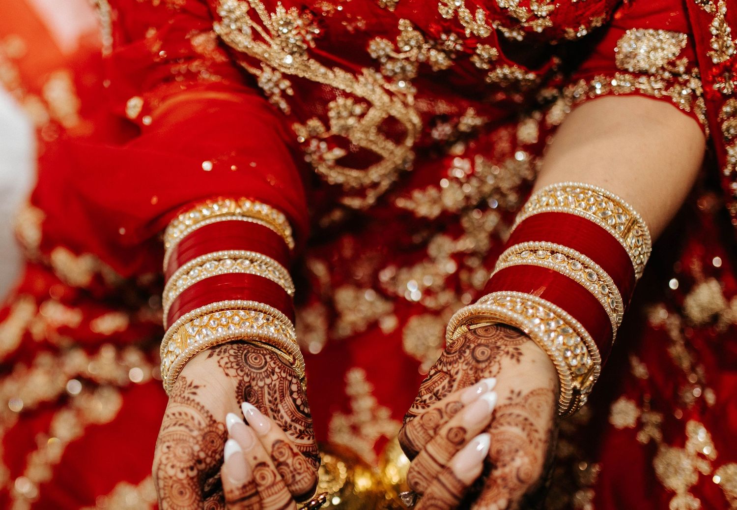 Hands decorated with intricate henna designs shown against vibrant red backdrop with gold decor details.
