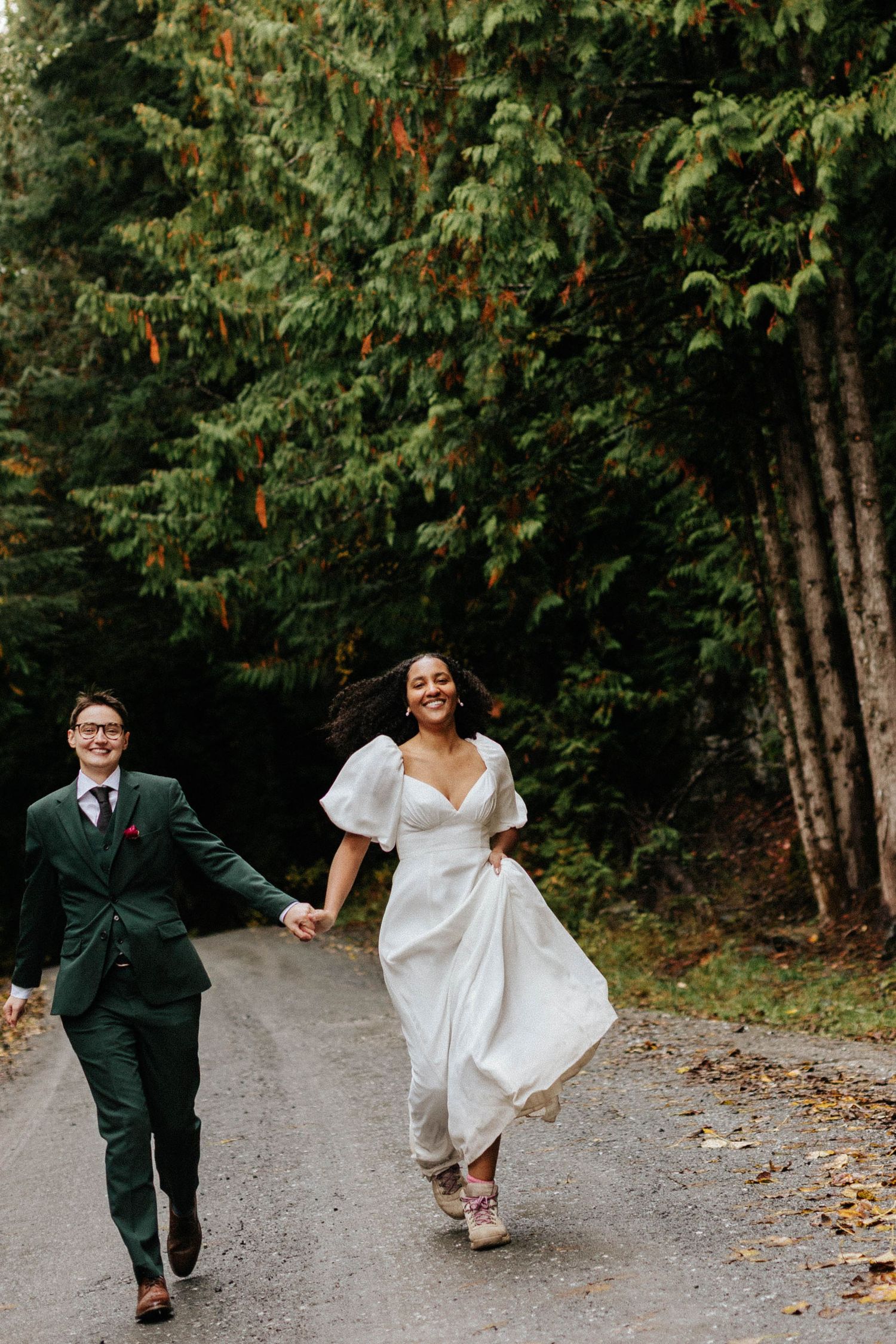 Joyful bride and groom in green suit run hand-in-hand down forested road during outdoor wedding.