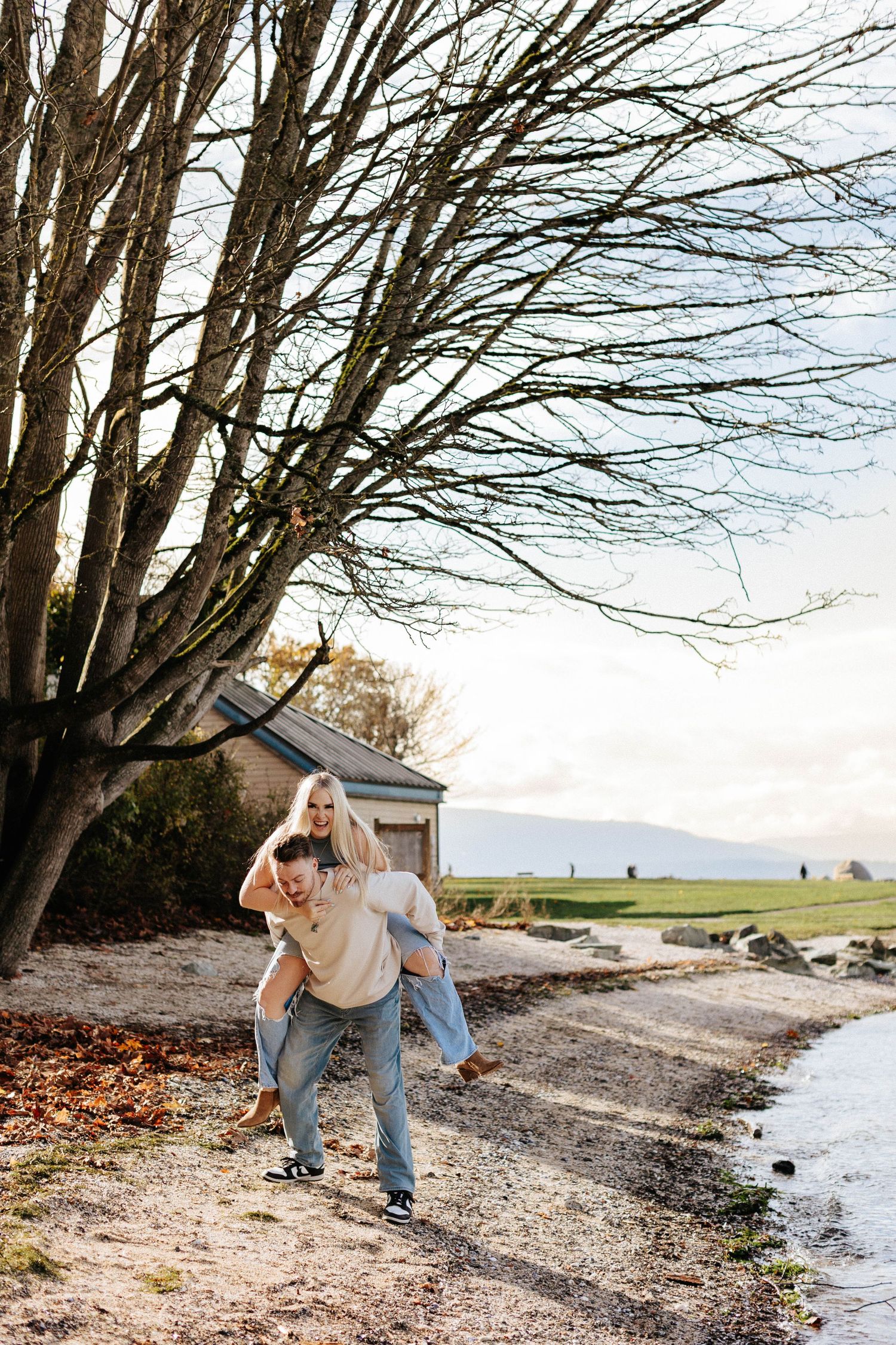Couple laughing as man gives woman a piggyback ride along a scenic waterfront path near bare trees.