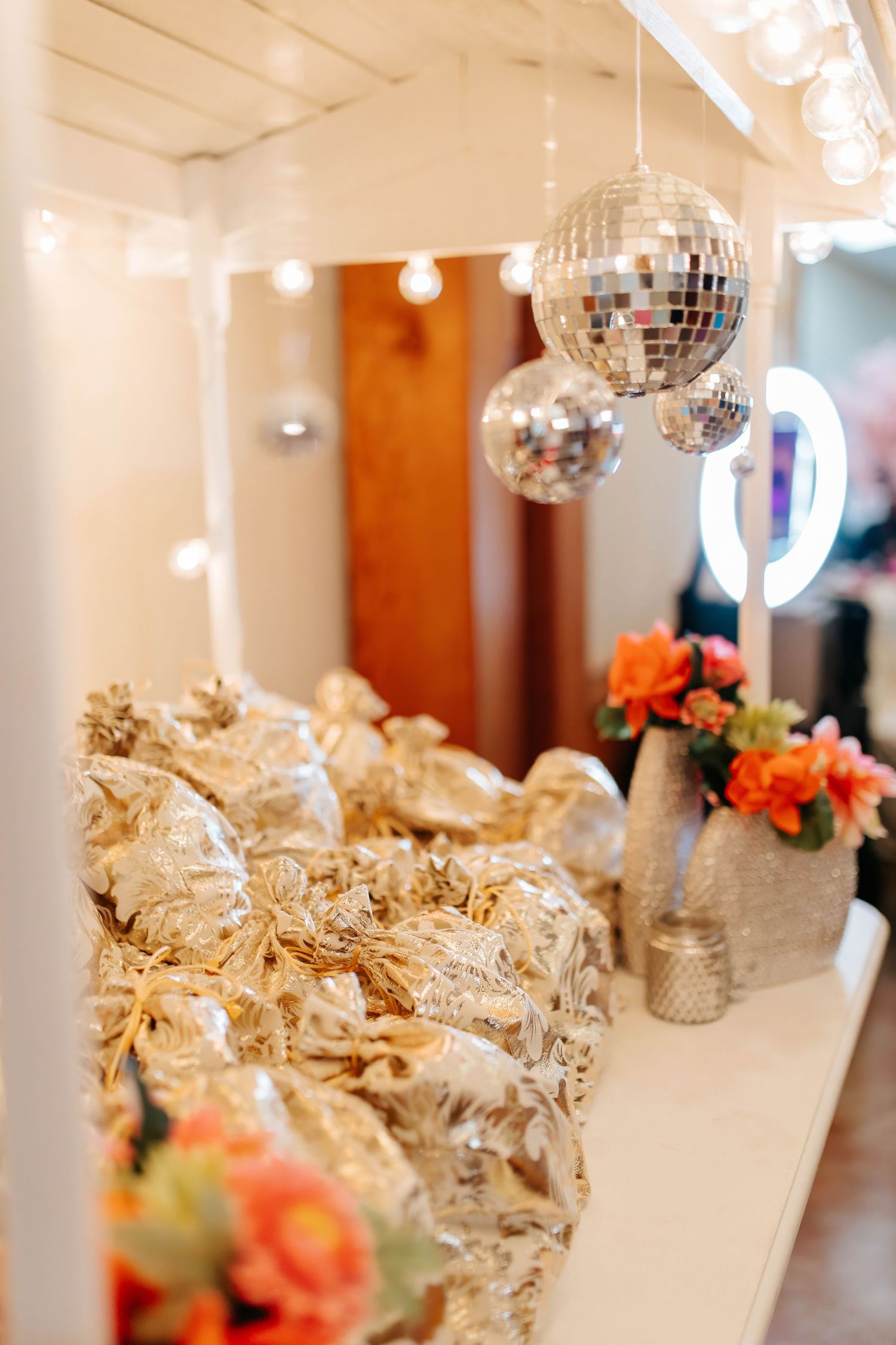 Silver disco ball hangs above elegant table setting with gold gift bags and coral flowers in party venue.