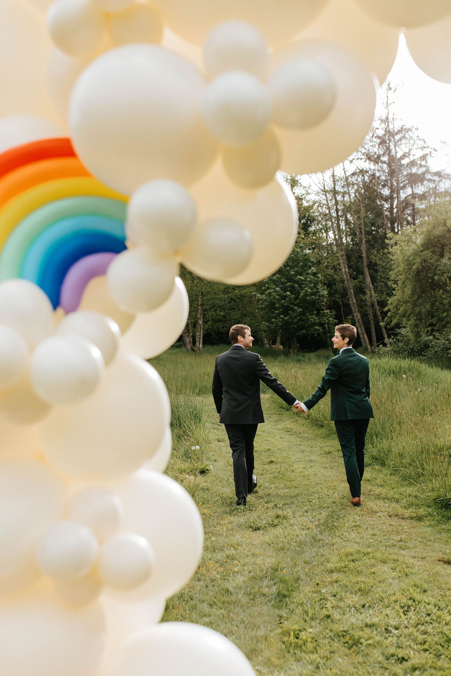 Two people in suits walk hand-in-hand on a grass path under a decorative rainbow and white balloon arch in nature.