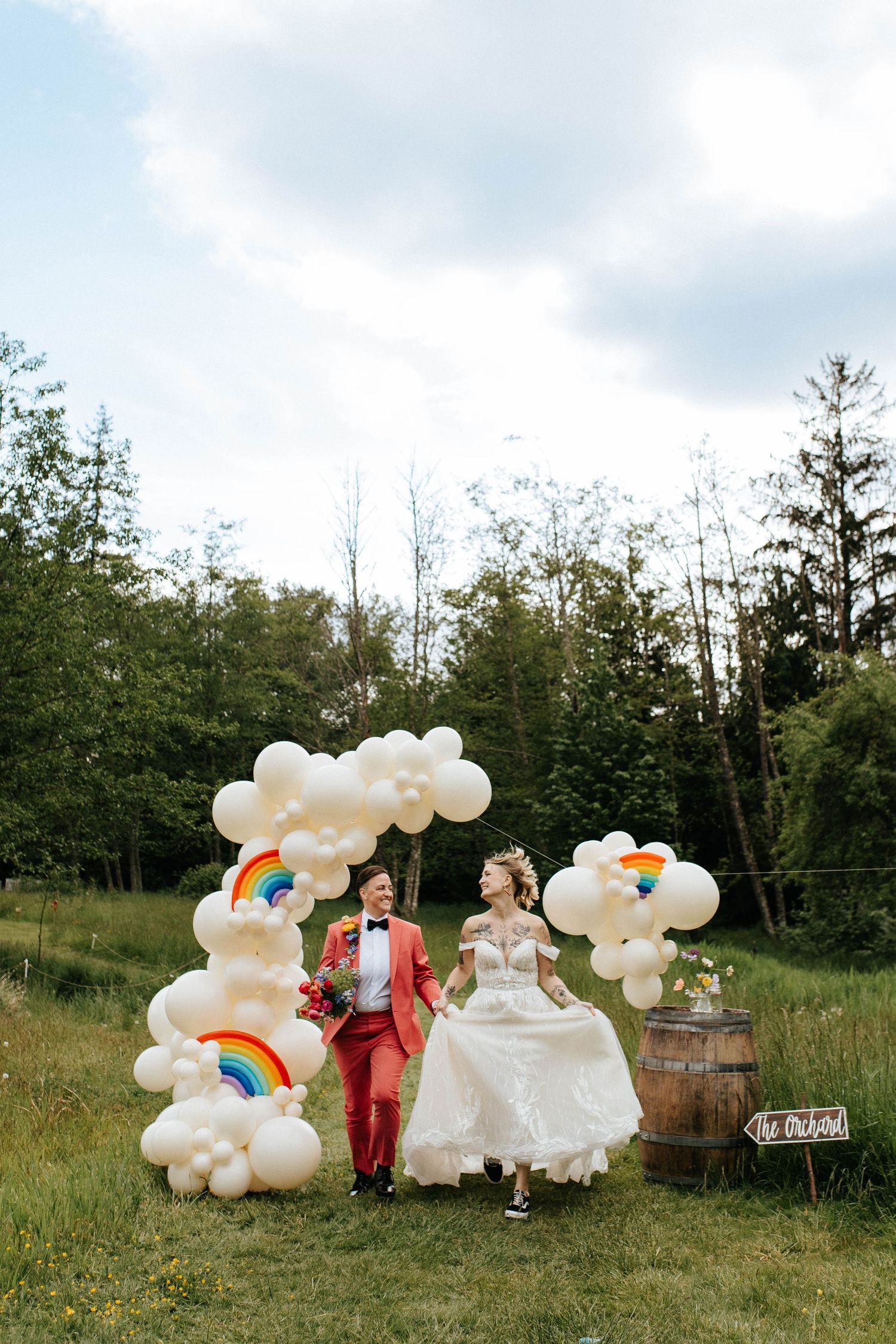 Outdoor wedding celebration with white balloon clouds and rainbow decorations in a rustic field setting.