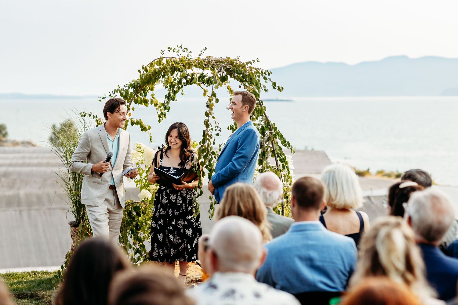 A scenic outdoor wedding ceremony takes place under a floral arch with mountains and water in the background.
