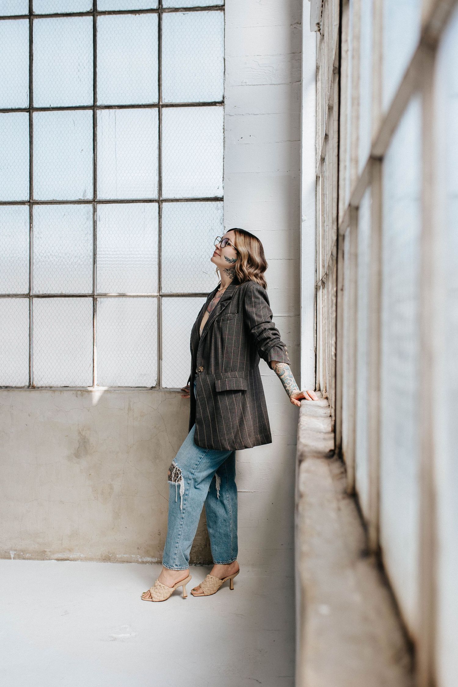 Woman in blazer and jeans poses by large industrial windows, looking up in a bright corridor.