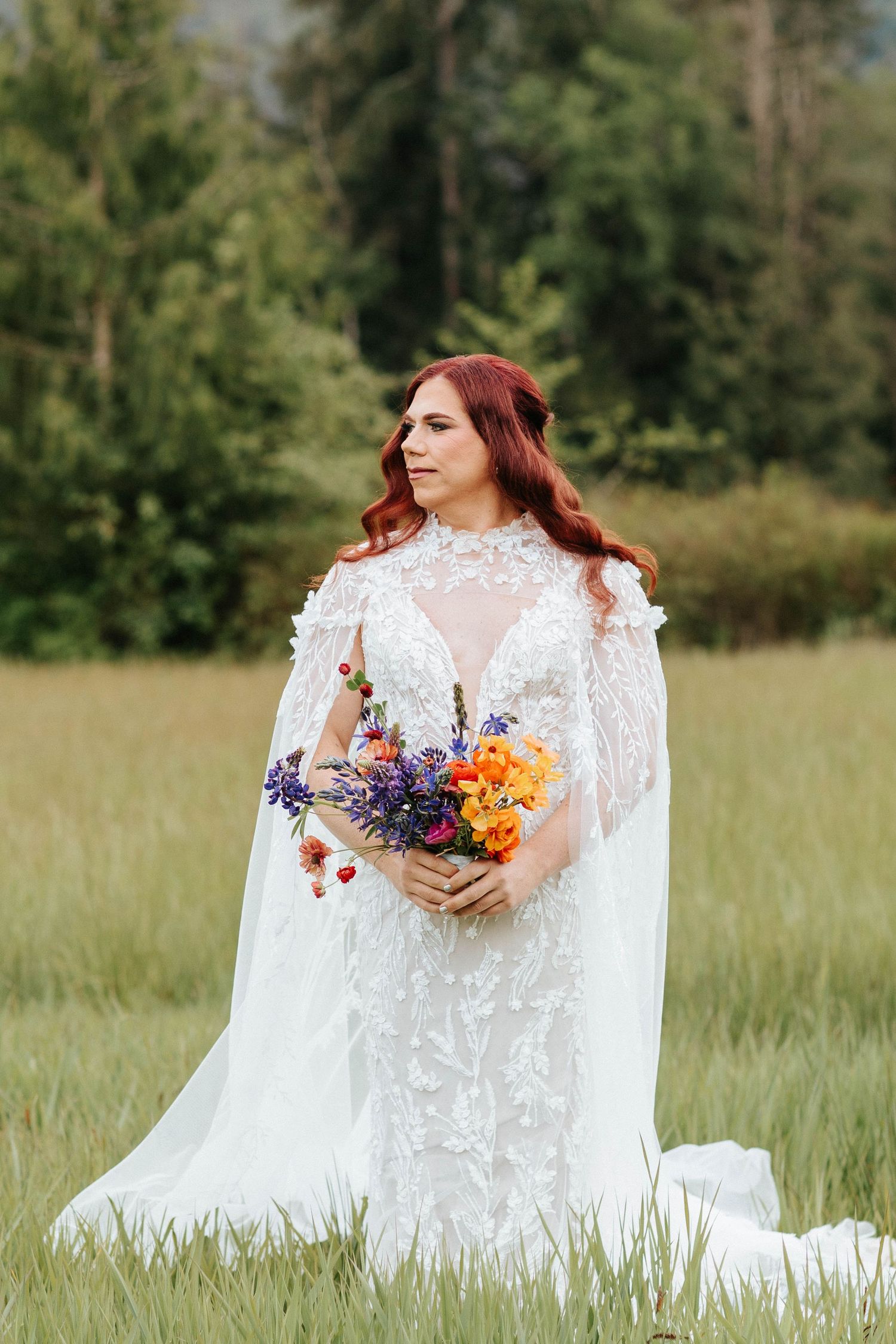 A bride in a white lace dress with long sleeves holds a vibrant purple and orange wildflower bouquet in a grassy meadow.