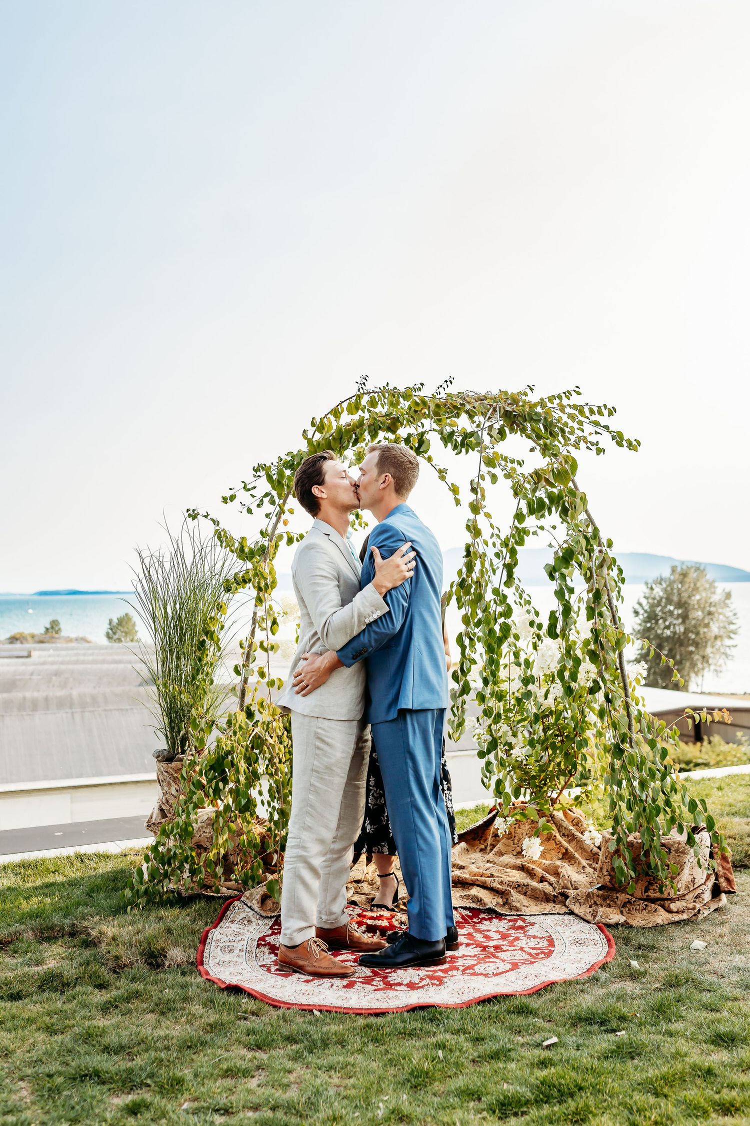 A couple embraces under a leafy green arch decorated with climbing vines on a grassy lawn by the waterfront.