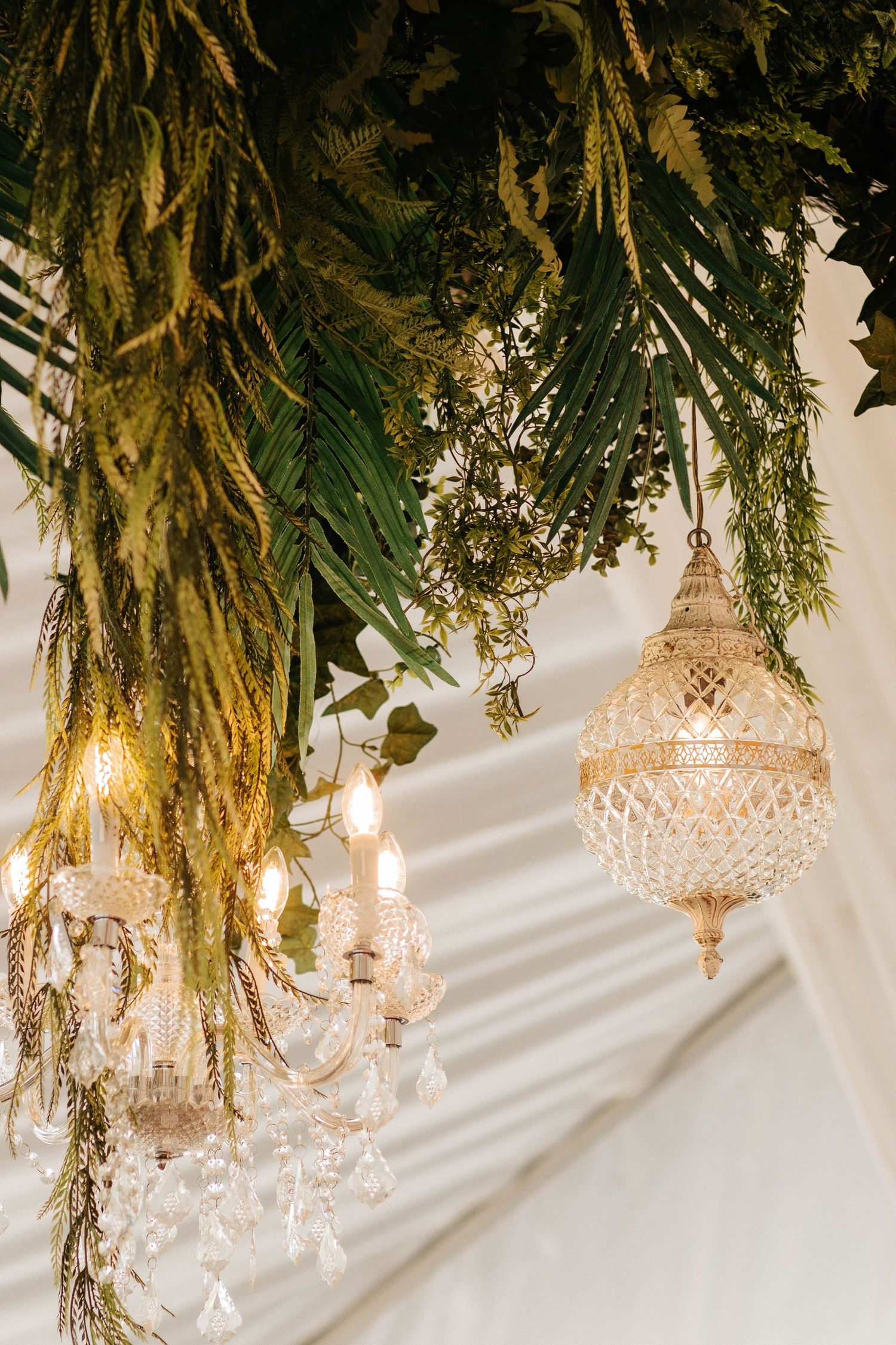 Elegant crystal chandeliers hang beneath lush cascading greenery and palm fronds at a luxury event venue.