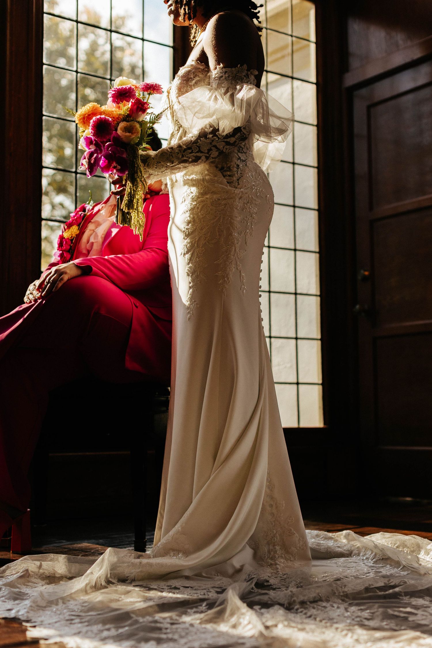 Bride in elegant lace gown stands by window with colorful bouquet beside seated groom in red suit.