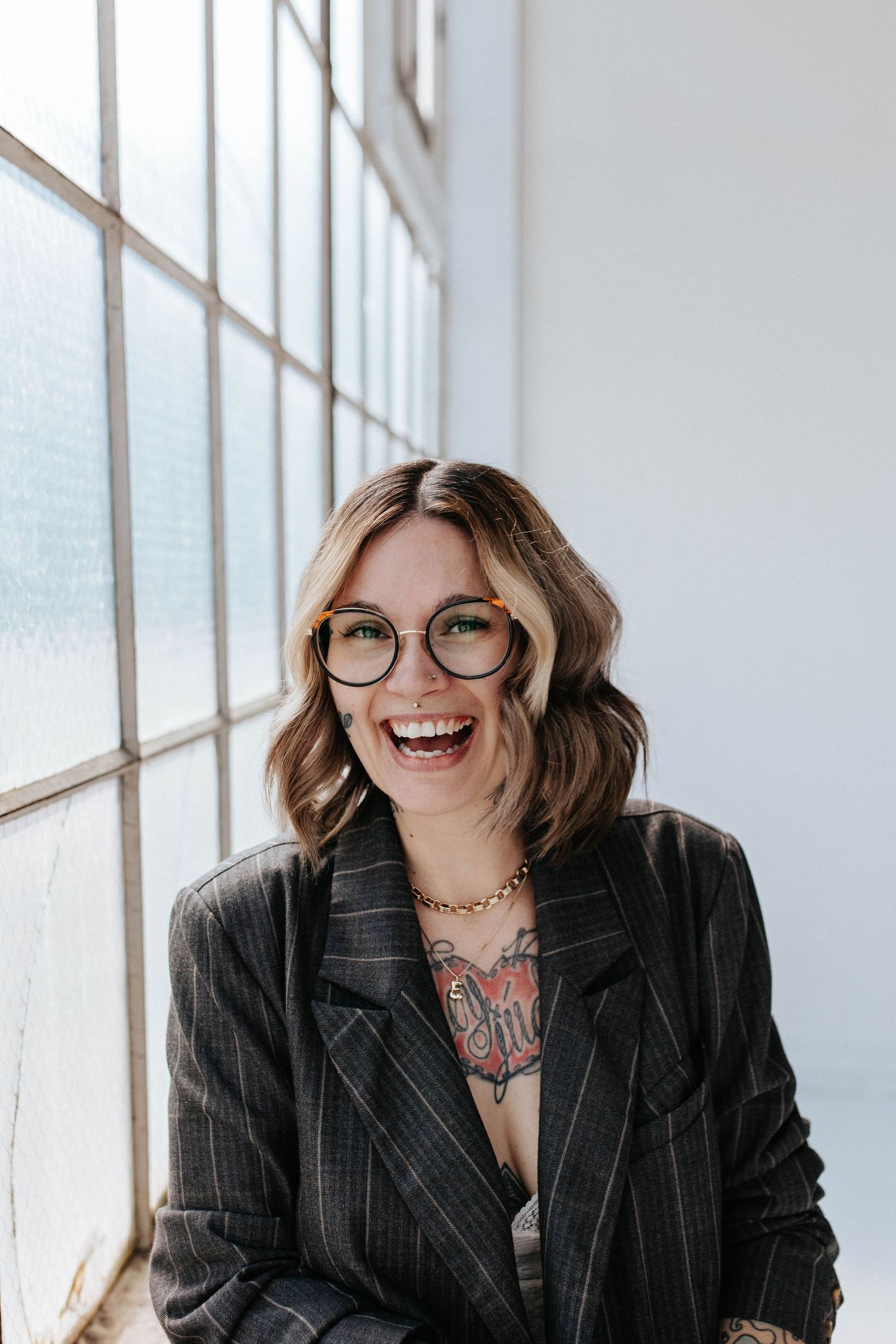 Smiling woman with glasses and tattoos wearing a dark blazer, laughing near a bright window.