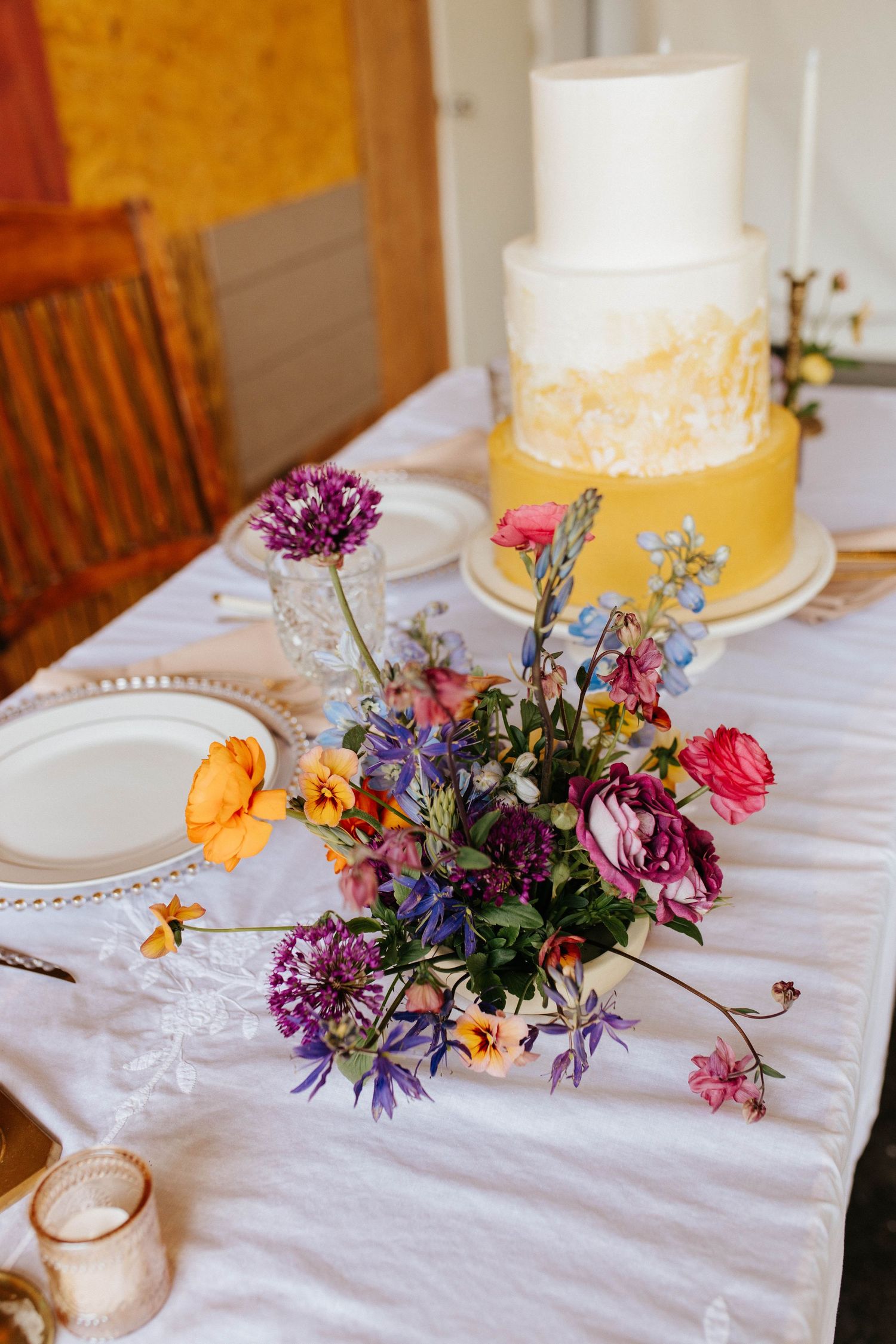 Yellow and white wedding cake displayed on table with vibrant purple and pink wildflower centerpiece arrangement.