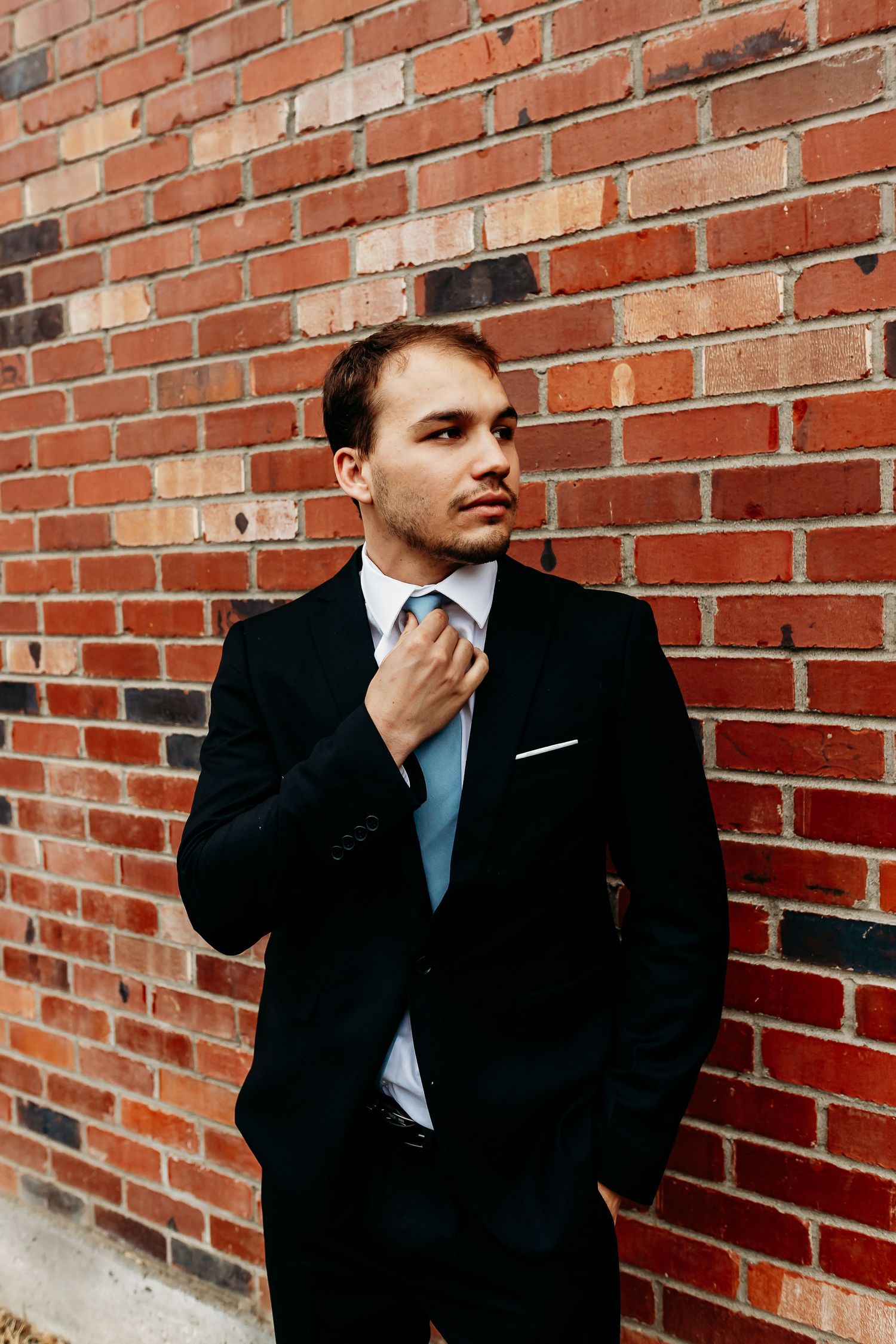 Professional in a dark suit and blue tie adjusts his necktie while standing against a rustic brick wall.