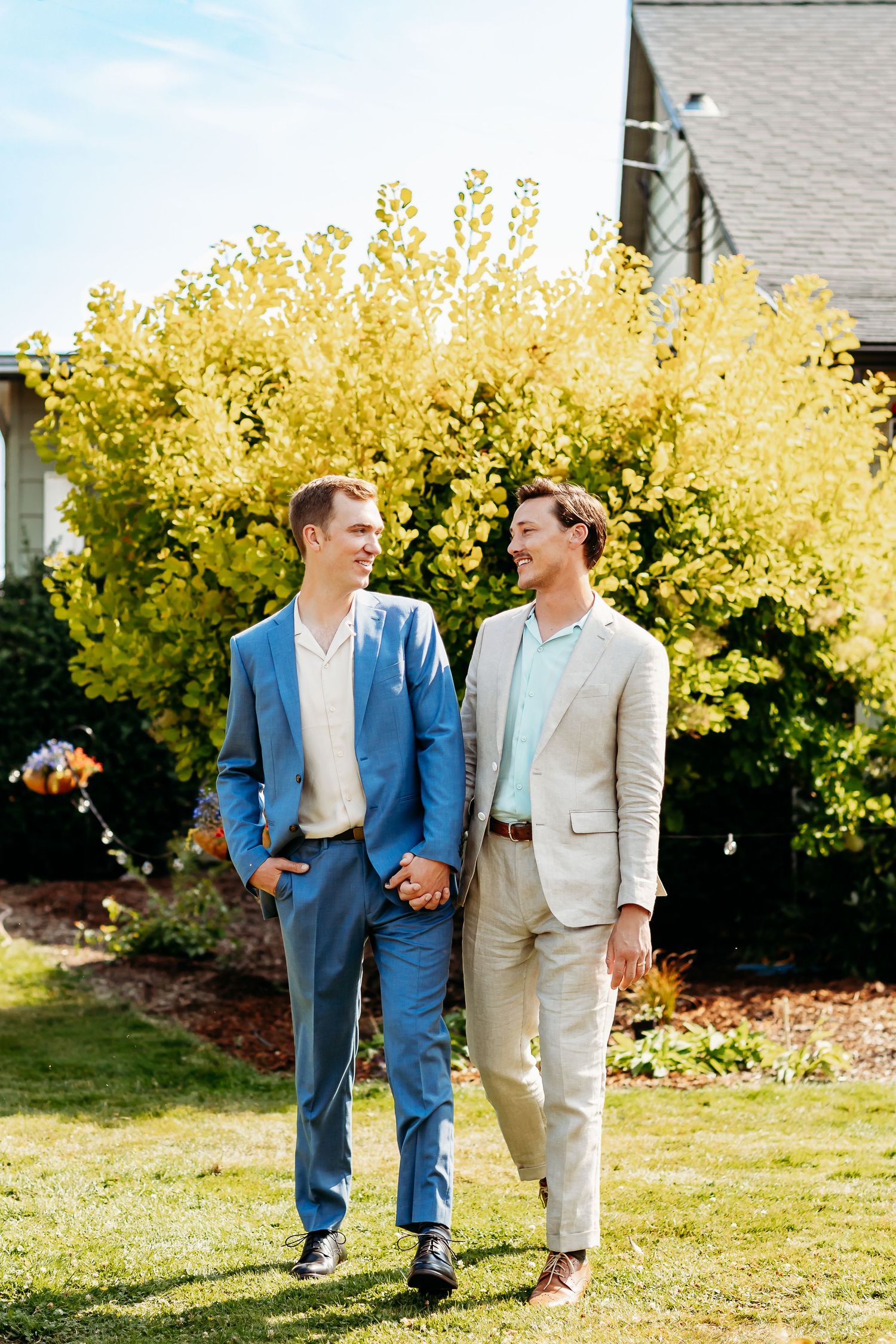 Two people in stylish suits walking together on green lawn with bright yellow forsythia bushes in background.