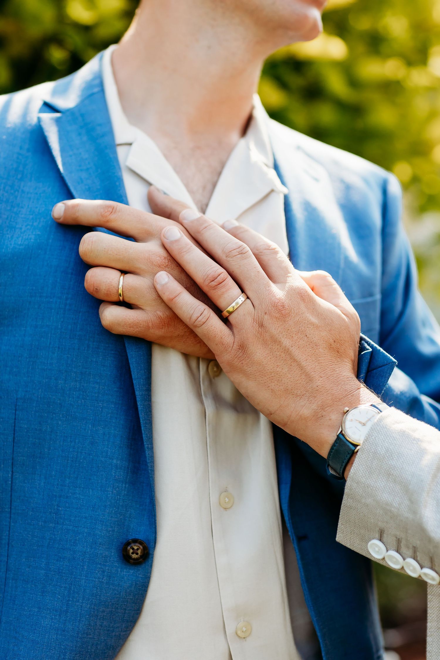 Close up of hands wearing wedding rings against a bright blue suit and white shirt in outdoor lighting.
