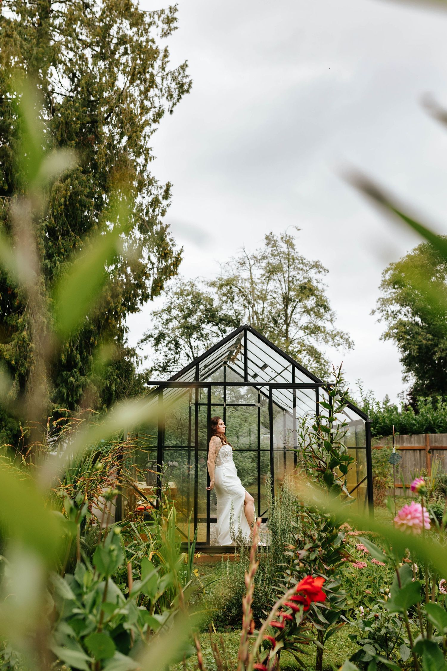 Bride in white gown stands beside a glass greenhouse surrounded by lush garden greenery and flowers.