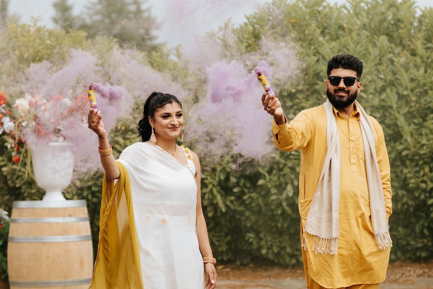 Group celebrates outdoor wedding ceremony with colorful smoke bombs in yellow and pink.