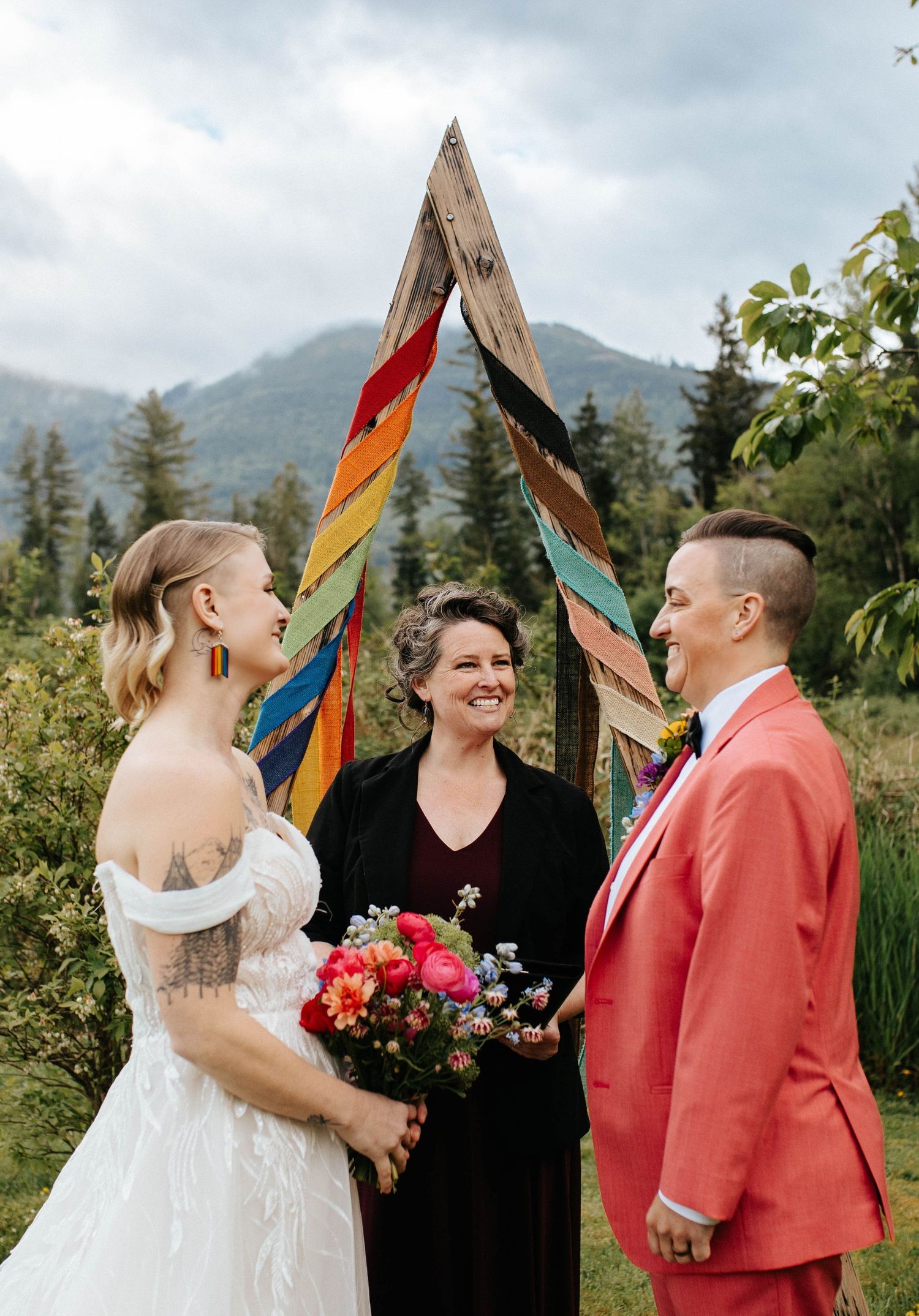 Wedding ceremony under a vibrant rainbow arch in a mountain setting with an officiant and couple in formal attire.