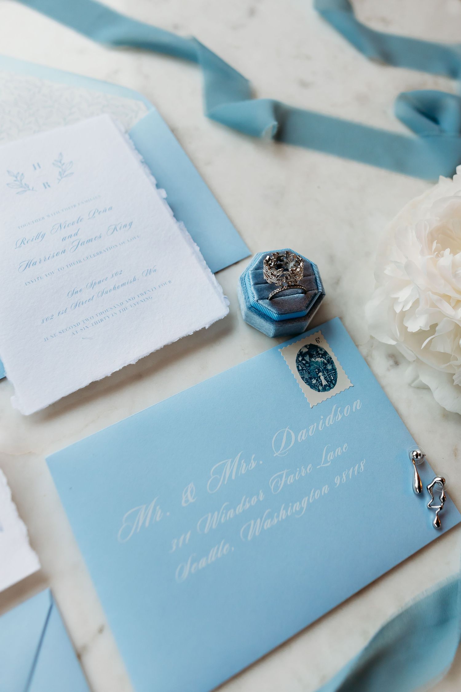 Light blue wedding invitation with white calligraphy text, silk ribbon, and decorative blue ring box on white surface.