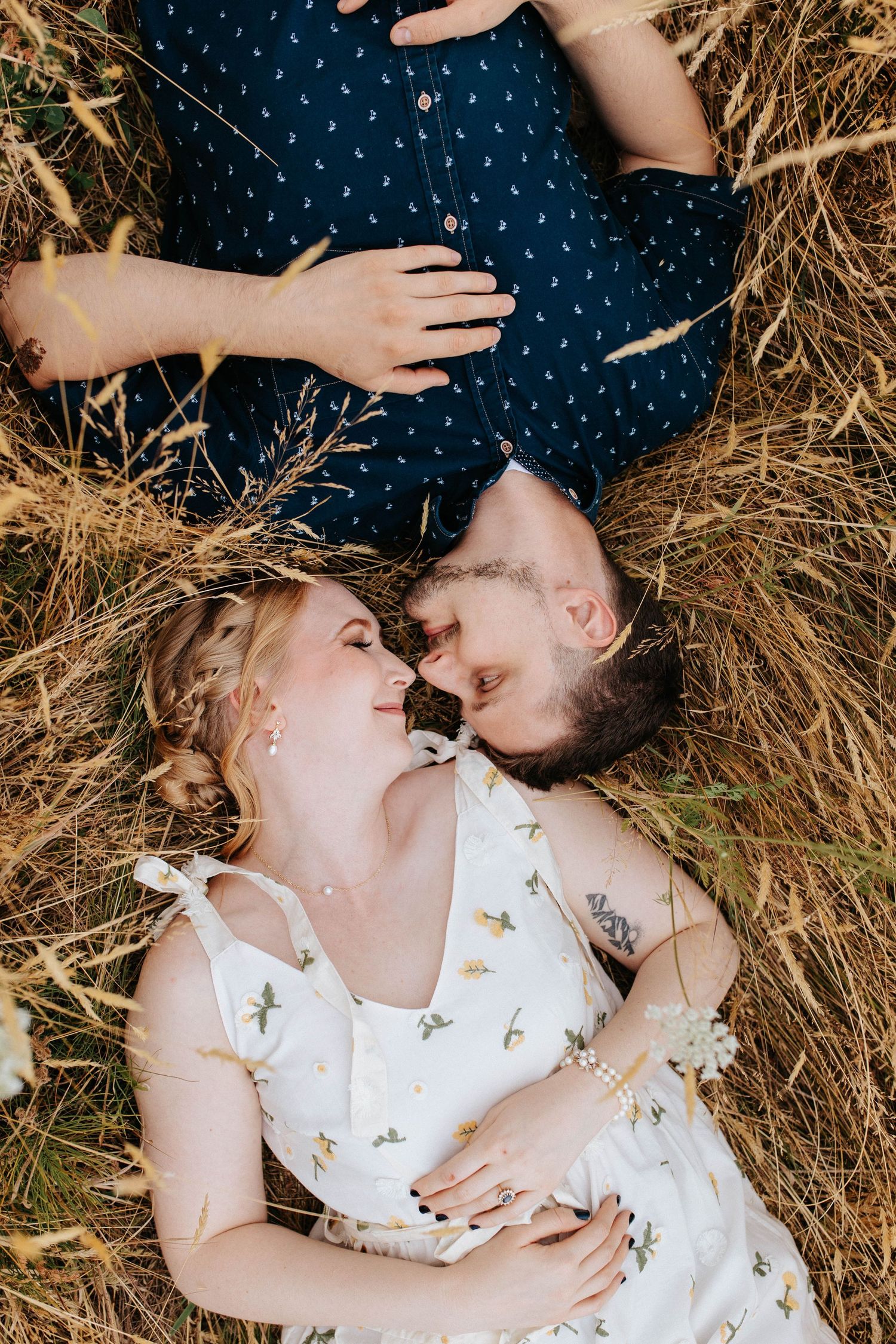 Couple lies in golden grass field, smiling at each other during a romantic outdoor engagement session.