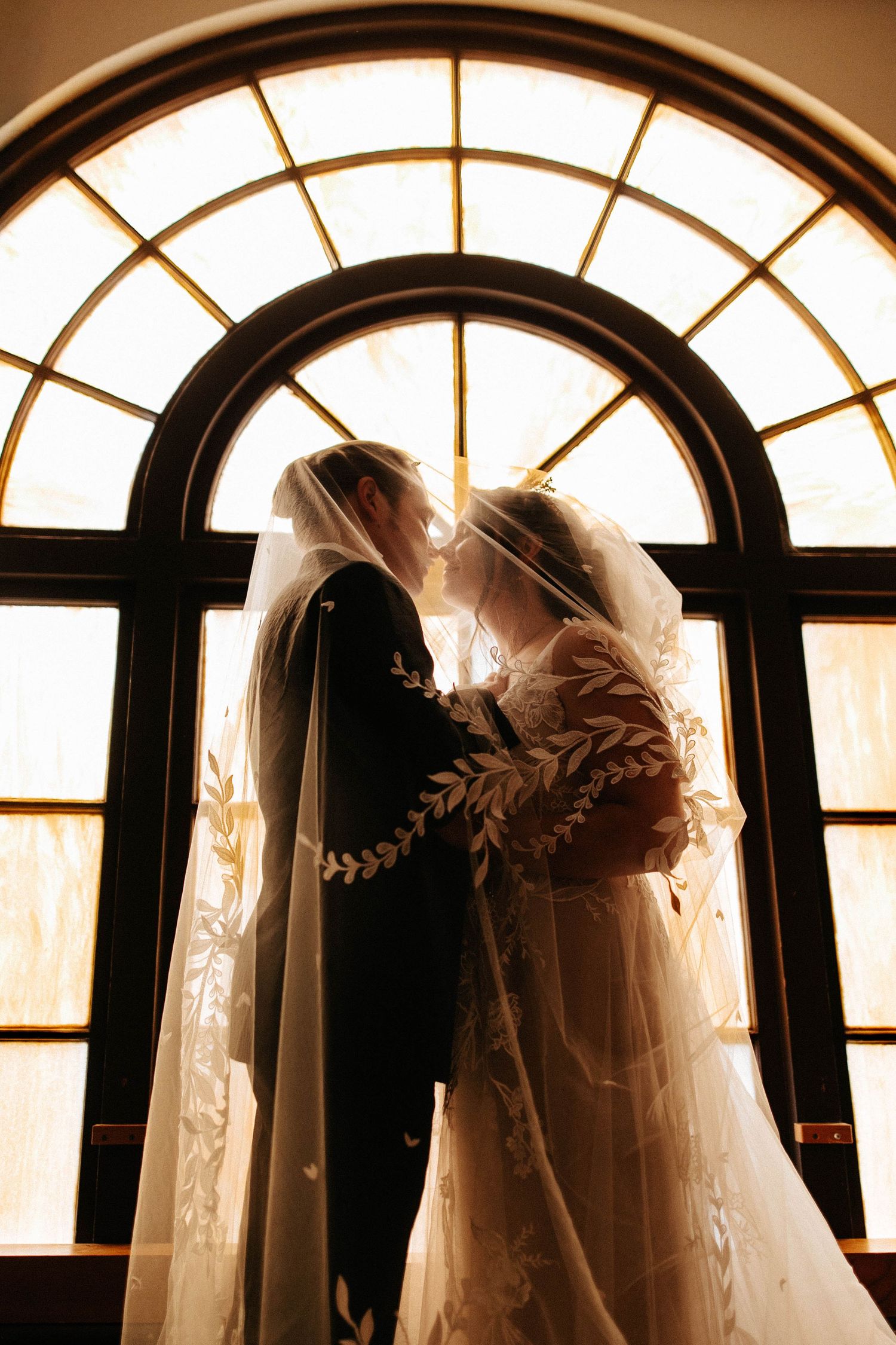 Bride and groom share an intimate kiss beneath an arched stained glass window with golden backlight.