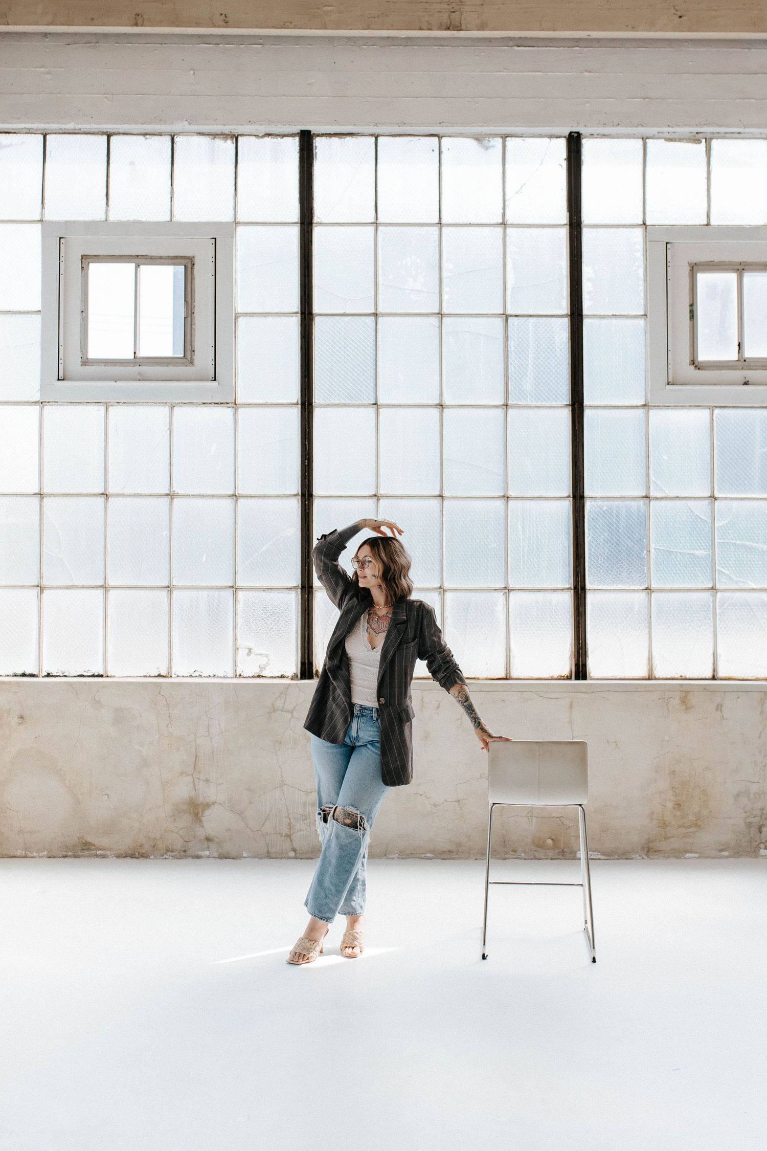 Woman in black leather jacket and ripped jeans posing by a white chair in a bright industrial loft space.