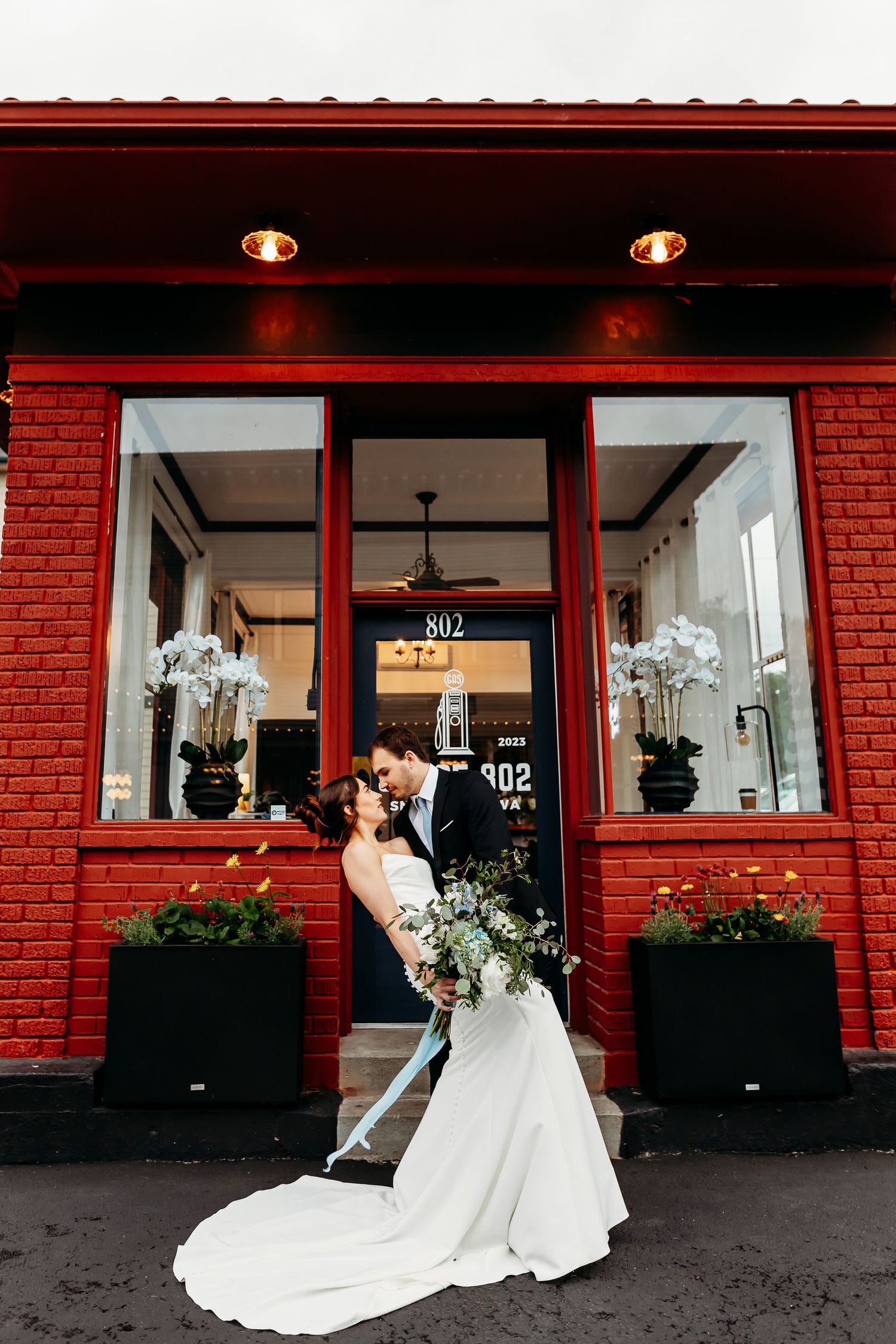 A couple embraces outside a red brick storefront with white flowers in black planters and white orchids in the window display.