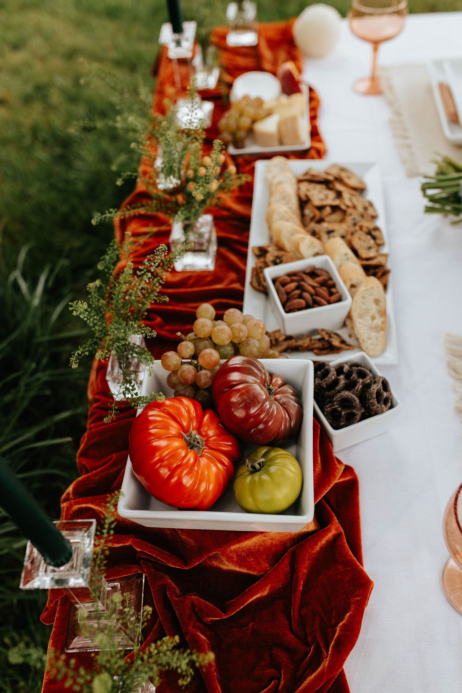 A fall table spread with heirloom tomatoes, mixed nuts, and snacks on a white tablecloth with red velvet runner.