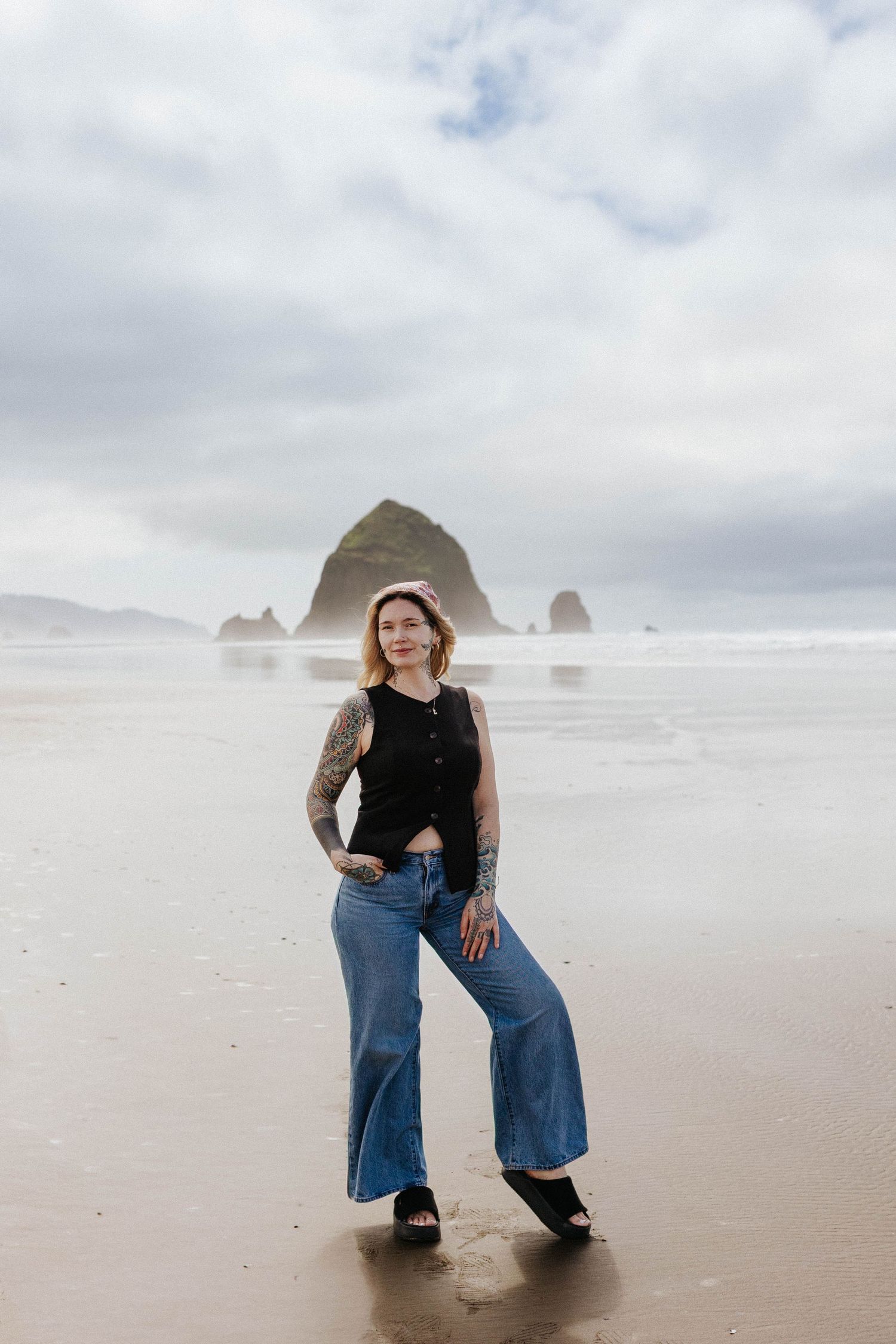 Tattooed woman in black sleeveless top and wide-leg jeans poses confidently on overcast Oregon beach.
