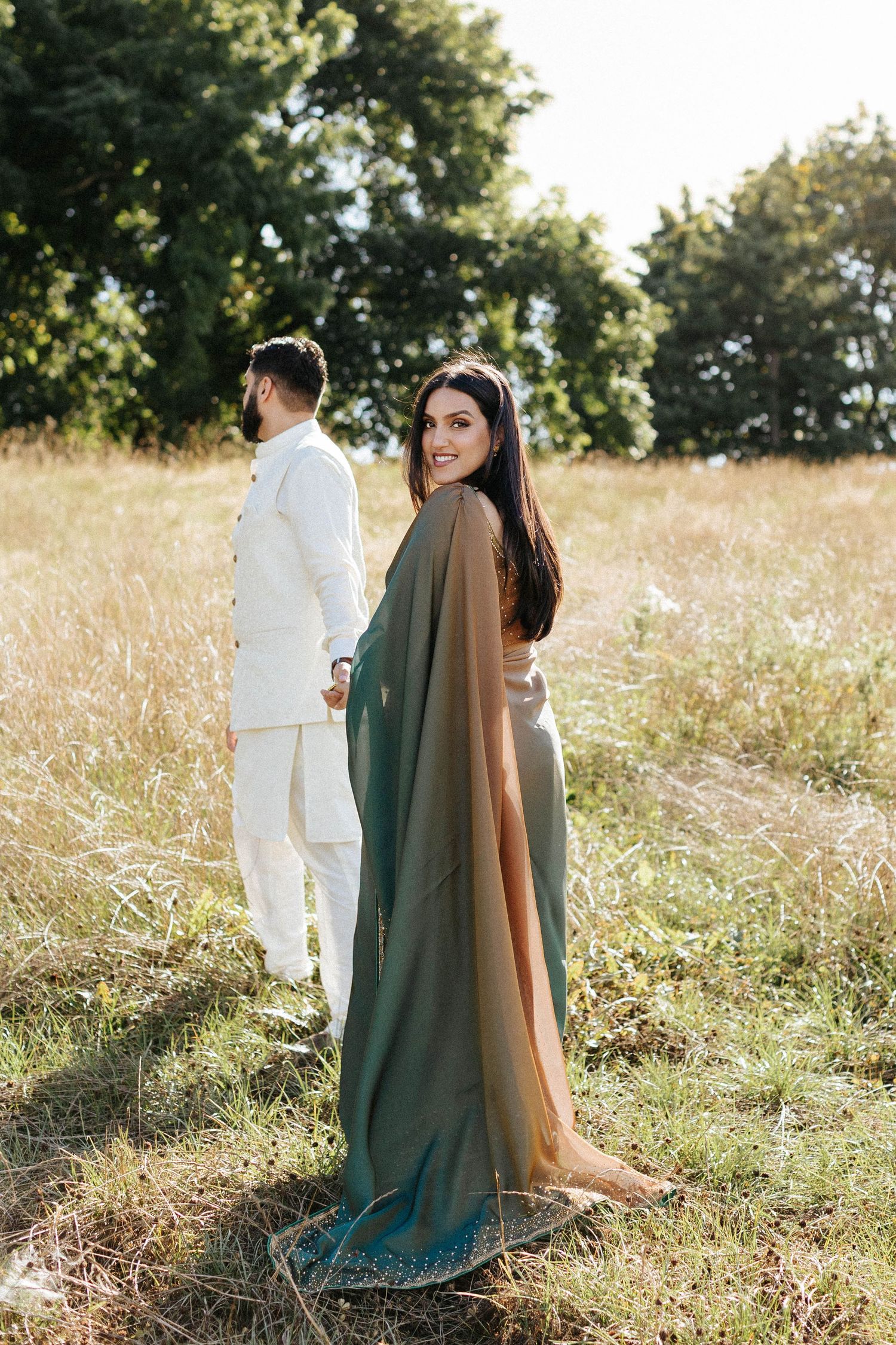 Bride in green and gold sari smiles back while holding groom's hand in a golden sunlit meadow.