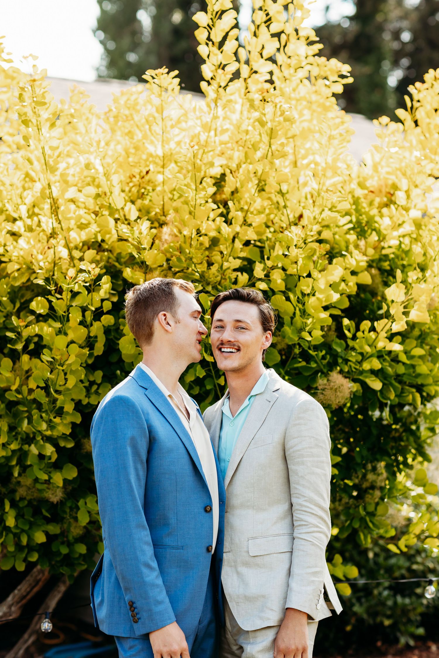 Happy couple in blue and beige blazers share a joyful moment in front of vibrant yellow foliage in a garden setting.