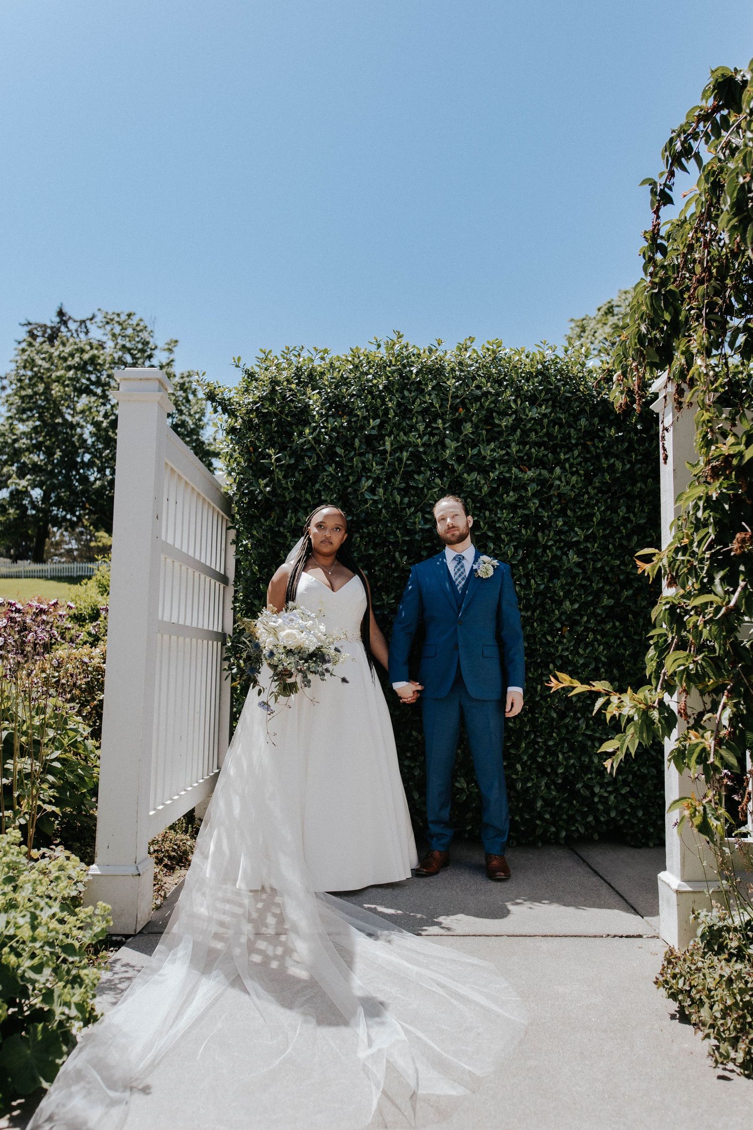 Bride in flowing white gown and groom in blue suit pose at a garden gate framed by lush green hedges.