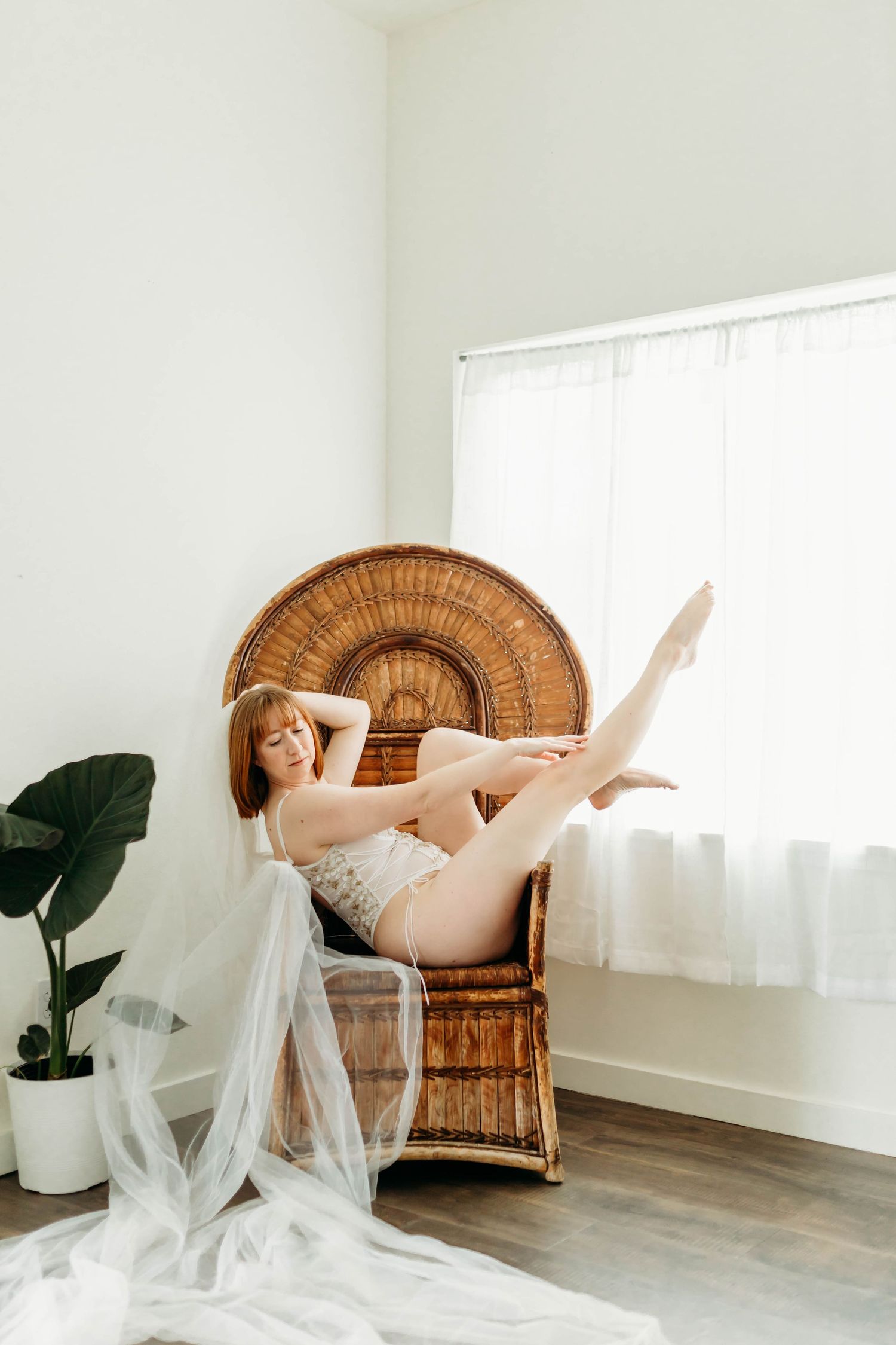Artistic boudoir photography with a rattan peacock chair, sheer veil, and potted plant in a bright minimal setting.