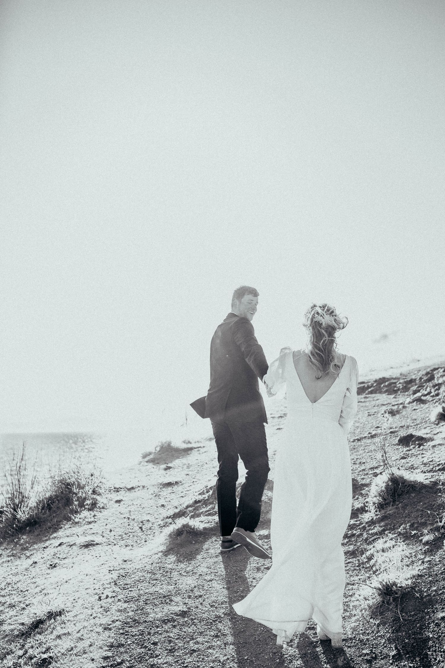 Black and white photo of bride and groom walking along a coastal hillside in the misty outdoors.