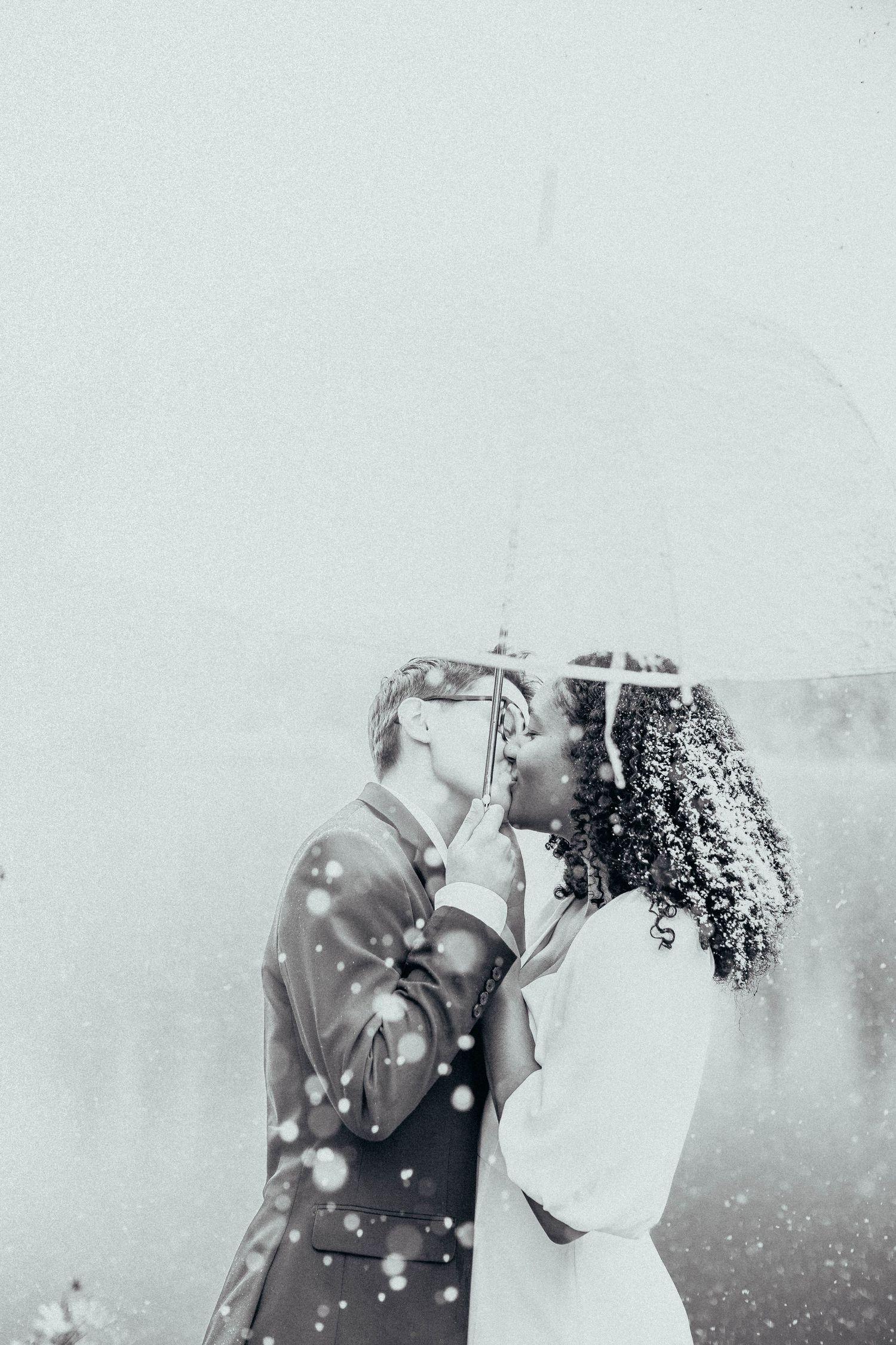 Couple sharing a romantic kiss under a clear umbrella in the rain, captured in soft black and white tones.
