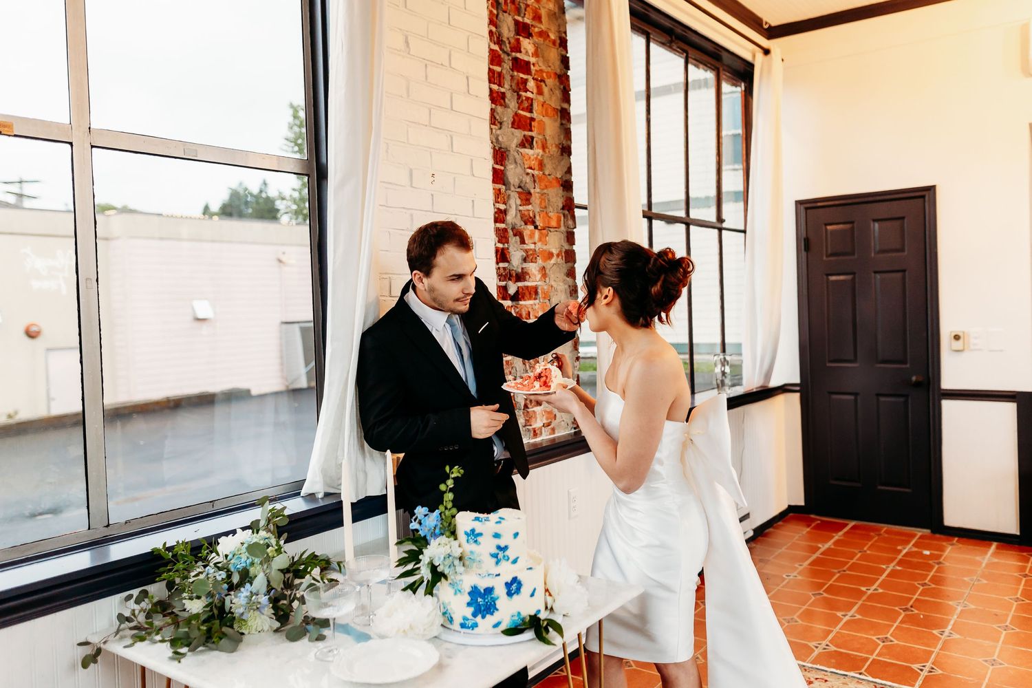 A bride and groom cut their wedding cake together in a rustic indoor venue with brick walls and large windows.