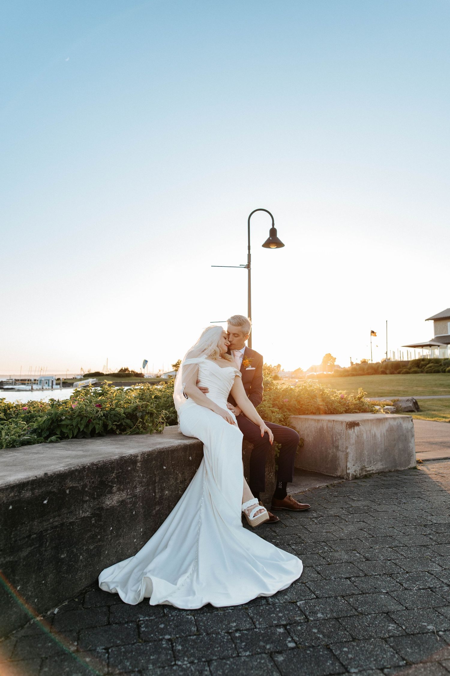 Bride and groom pose at sunset near a waterfront, her mermaid gown trailing gracefully on stone steps.