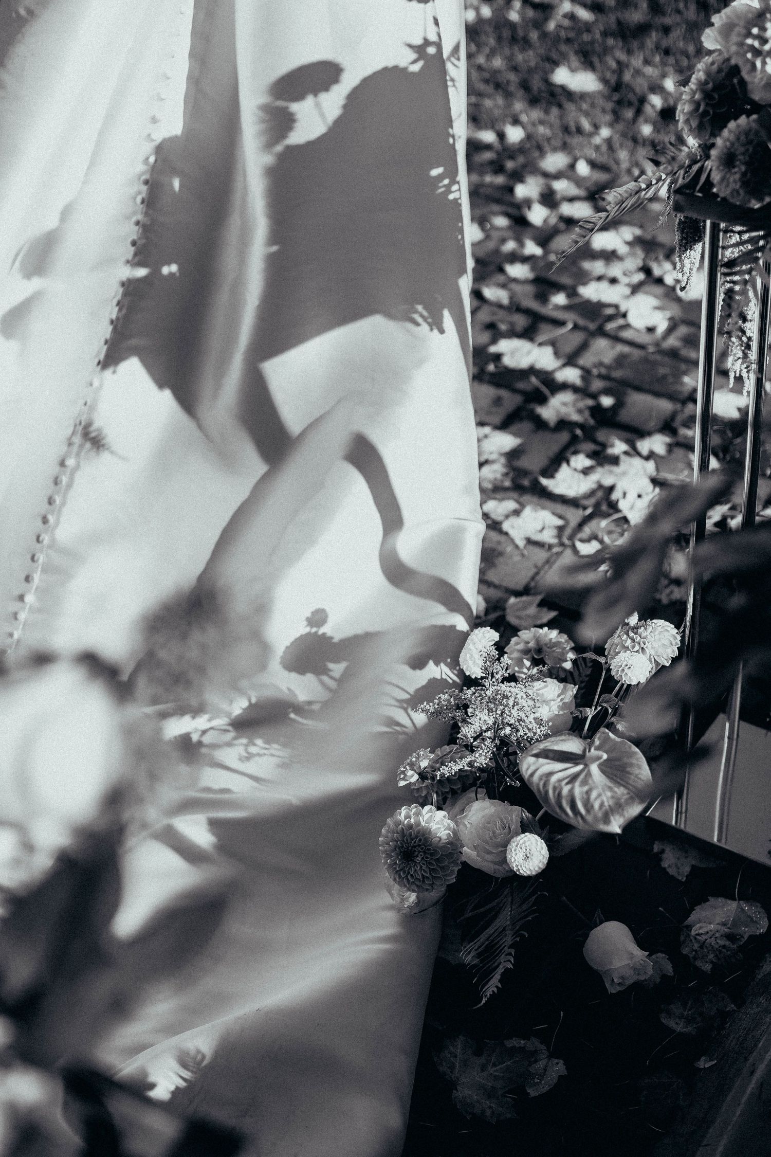 Black and white artistic shot of a wedding dress hem with a floral bouquet on cobblestone steps.