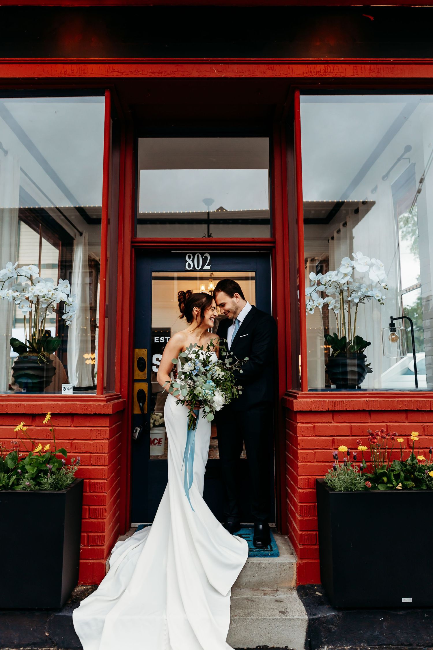 A romantic wedding photo outside a building with a striking red storefront and flowers, featuring a long flowing white gown.