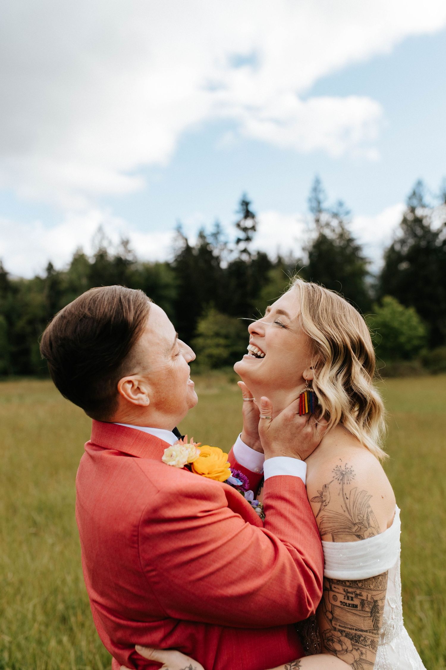 Two people share joyful laughter while embracing in a sunlit field with evergreen trees in the background.