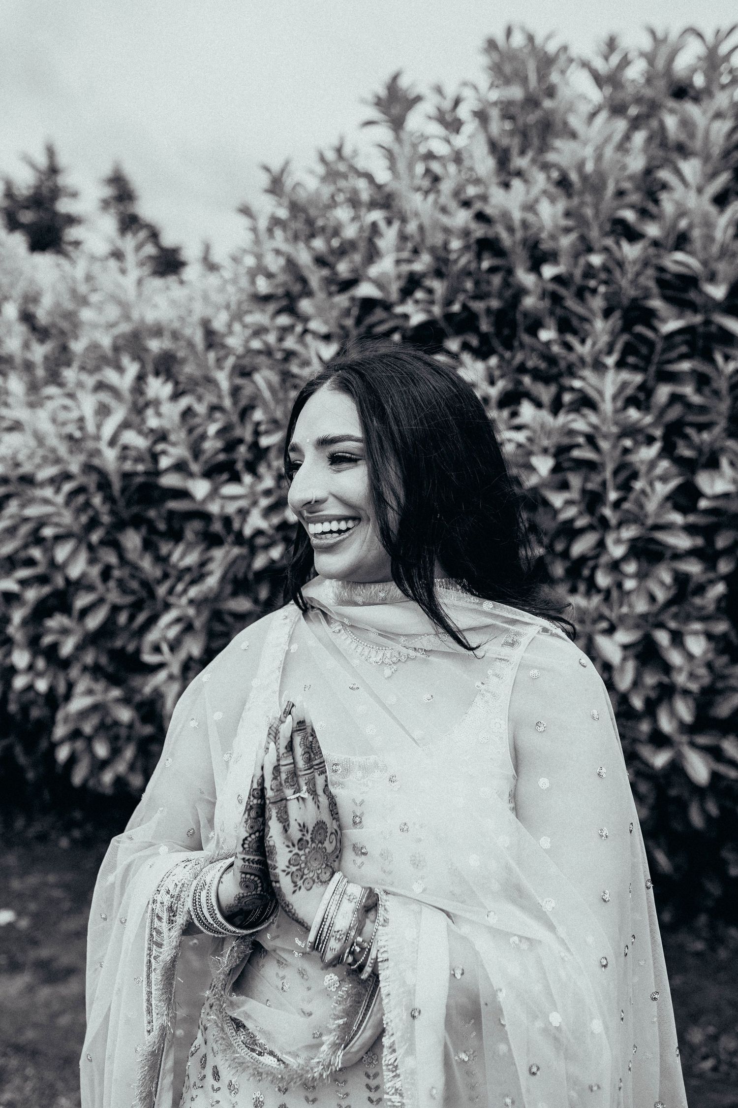 Black and white portrait of someone in traditional South Asian attire with henna-decorated hands smiling outdoors.