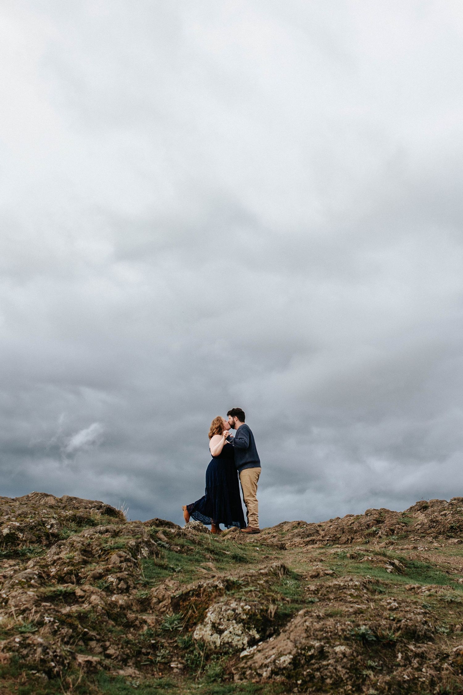 Couple embraces on a rocky hilltop beneath dramatic stormy clouds during an outdoor engagement session.