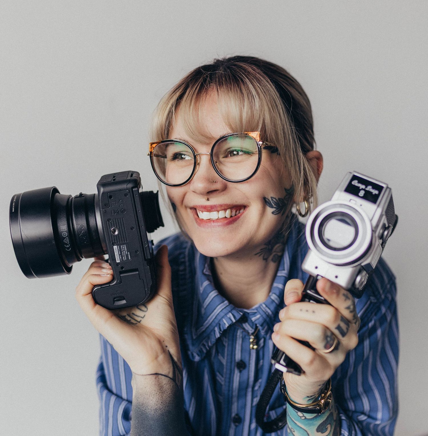 Smiling tattooed woman with glasses holds two cameras against a light gray studio background.