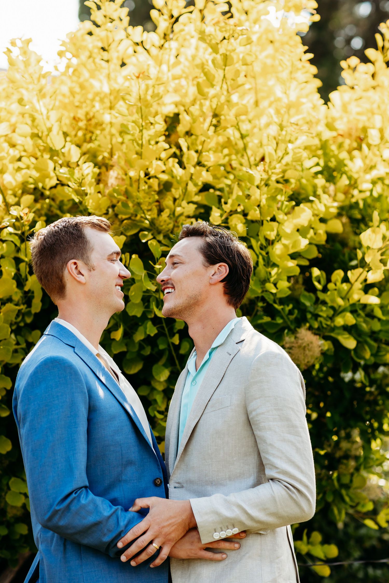 A couple in blazers share a joyful moment together against blooming yellow flowers on a sunny day.
