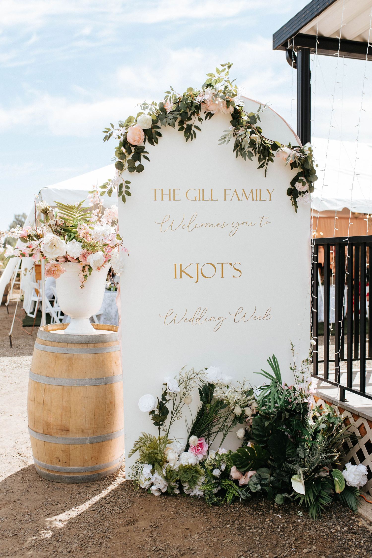 Elegant wedding welcome sign with gold lettering and floral decorations next to a rustic wine barrel at an outdoor venue.