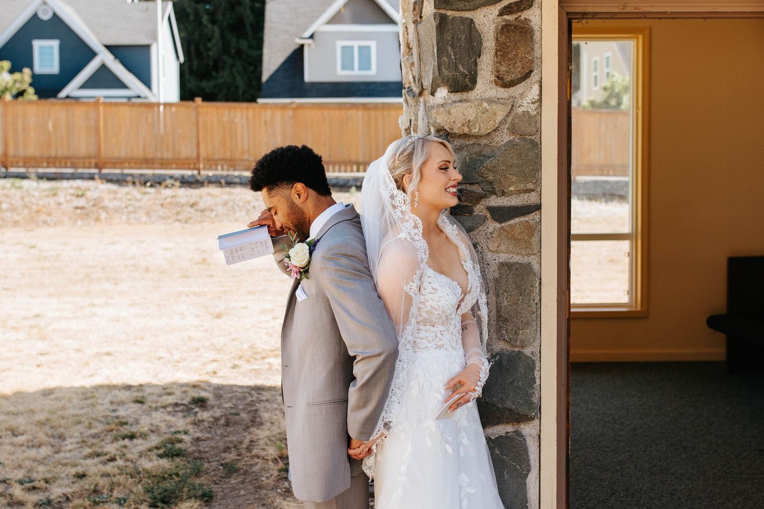 Bride and groom share a sweet first look moment, standing back-to-back against a stone wall outdoors.