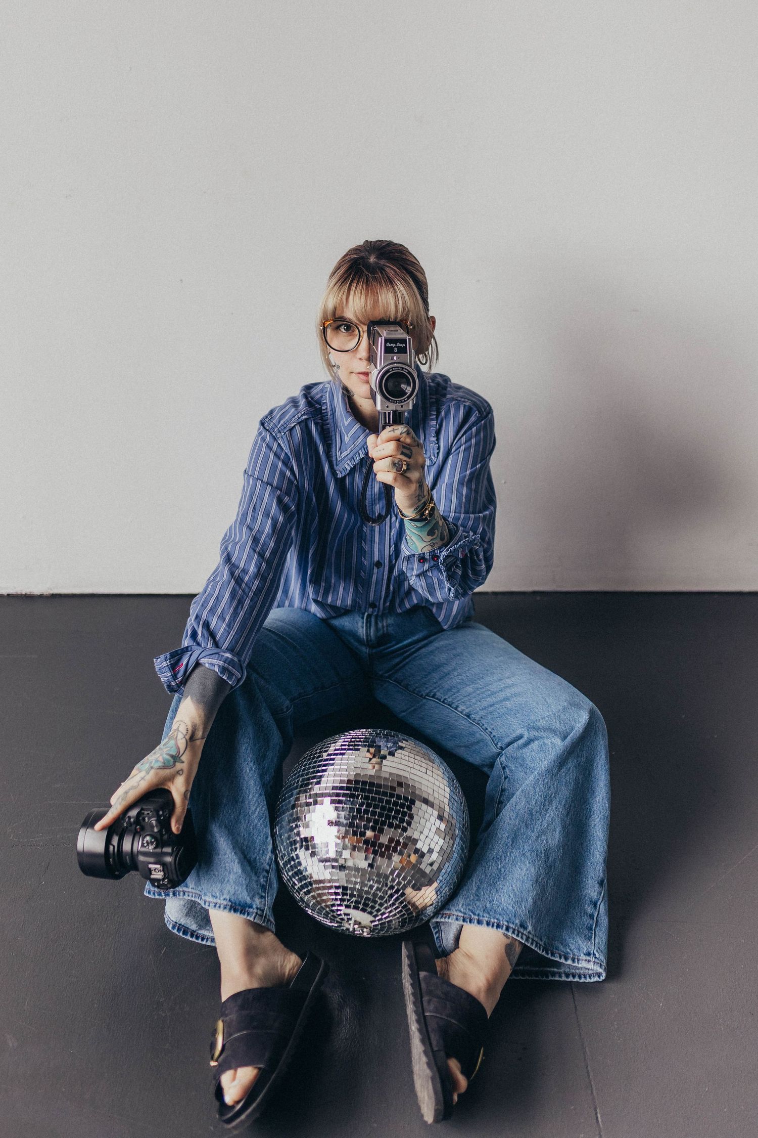 Tattooed woman in blue striped shirt sits on disco ball holding a vintage camera in studio.
