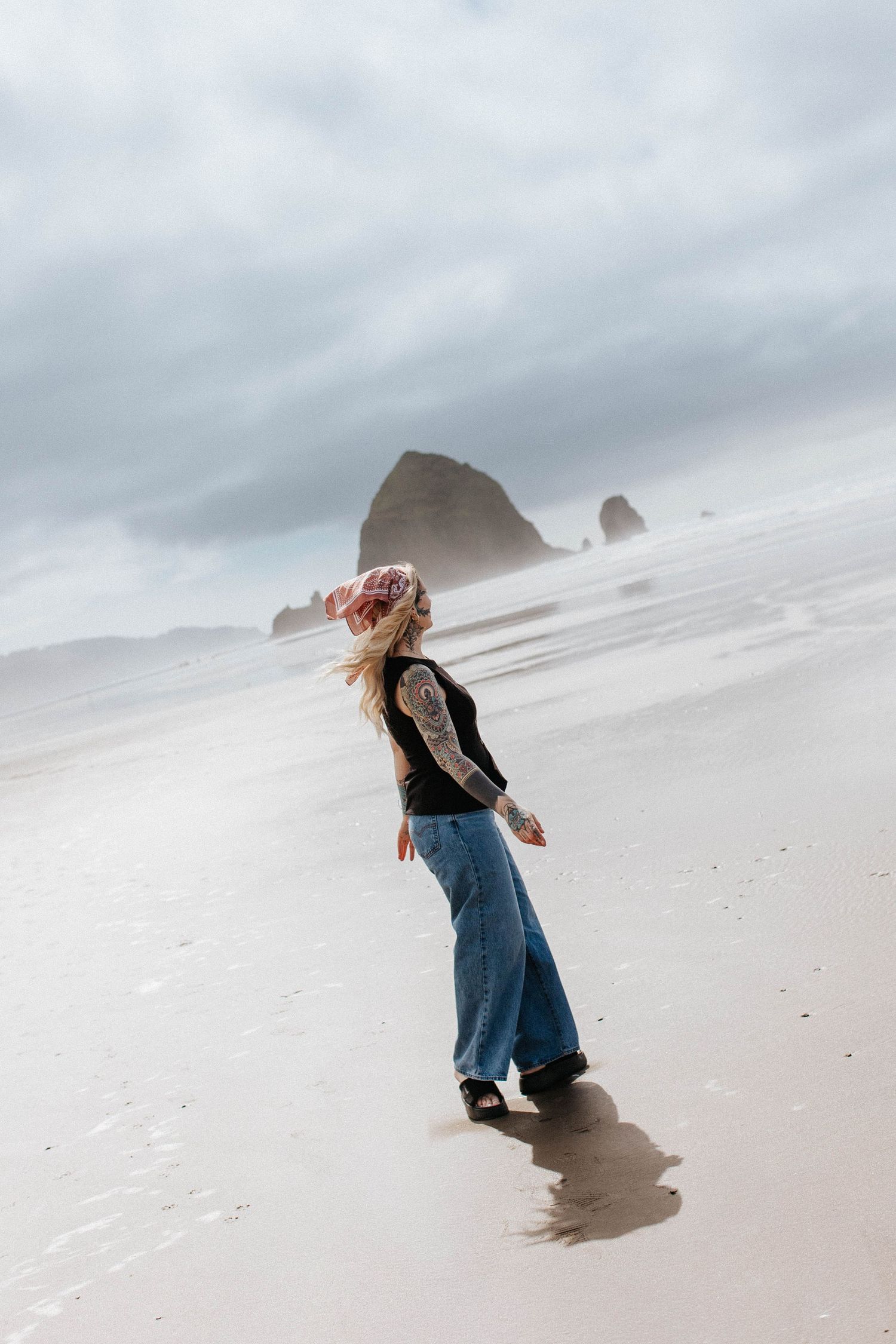 Tattooed woman in black top and wide-leg jeans walks on sandy beach with Haystack Rock behind her.